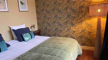 A neatly made bed with a green quilt, a wall with a bird pattern wallpaper, and a lamp on a side table.