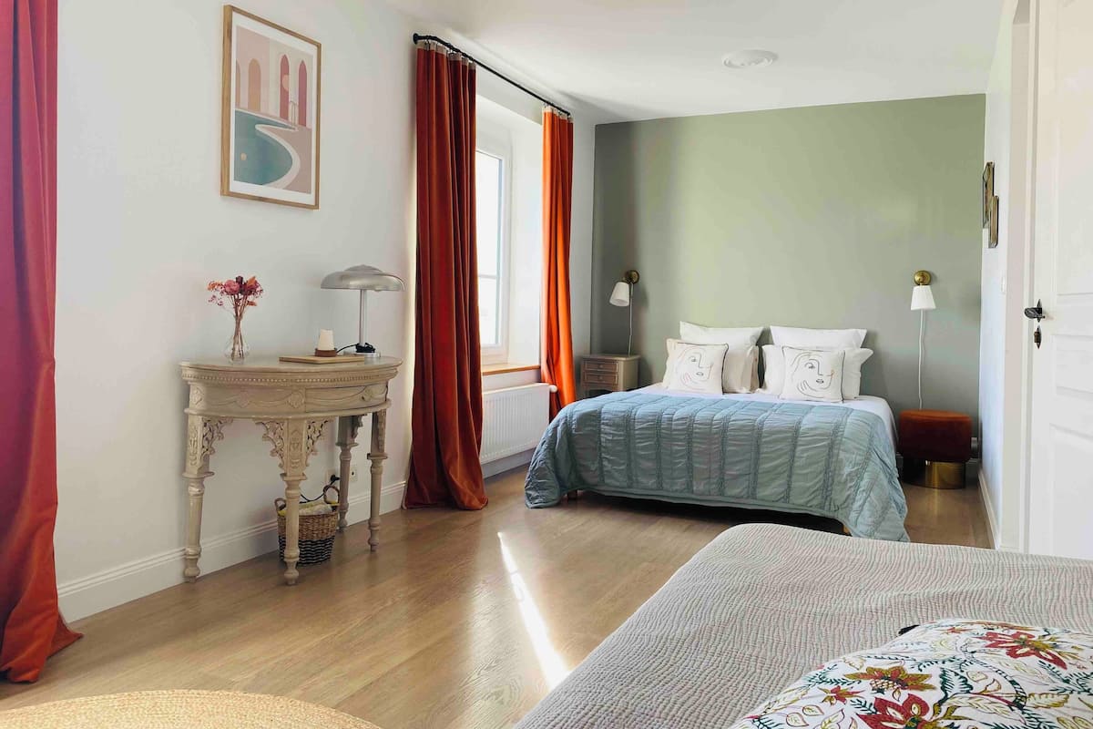 Une chambre à coucher comprenant un lit, des tables de chevet, une commode, une fenêtre avec des rideaux et une porte.