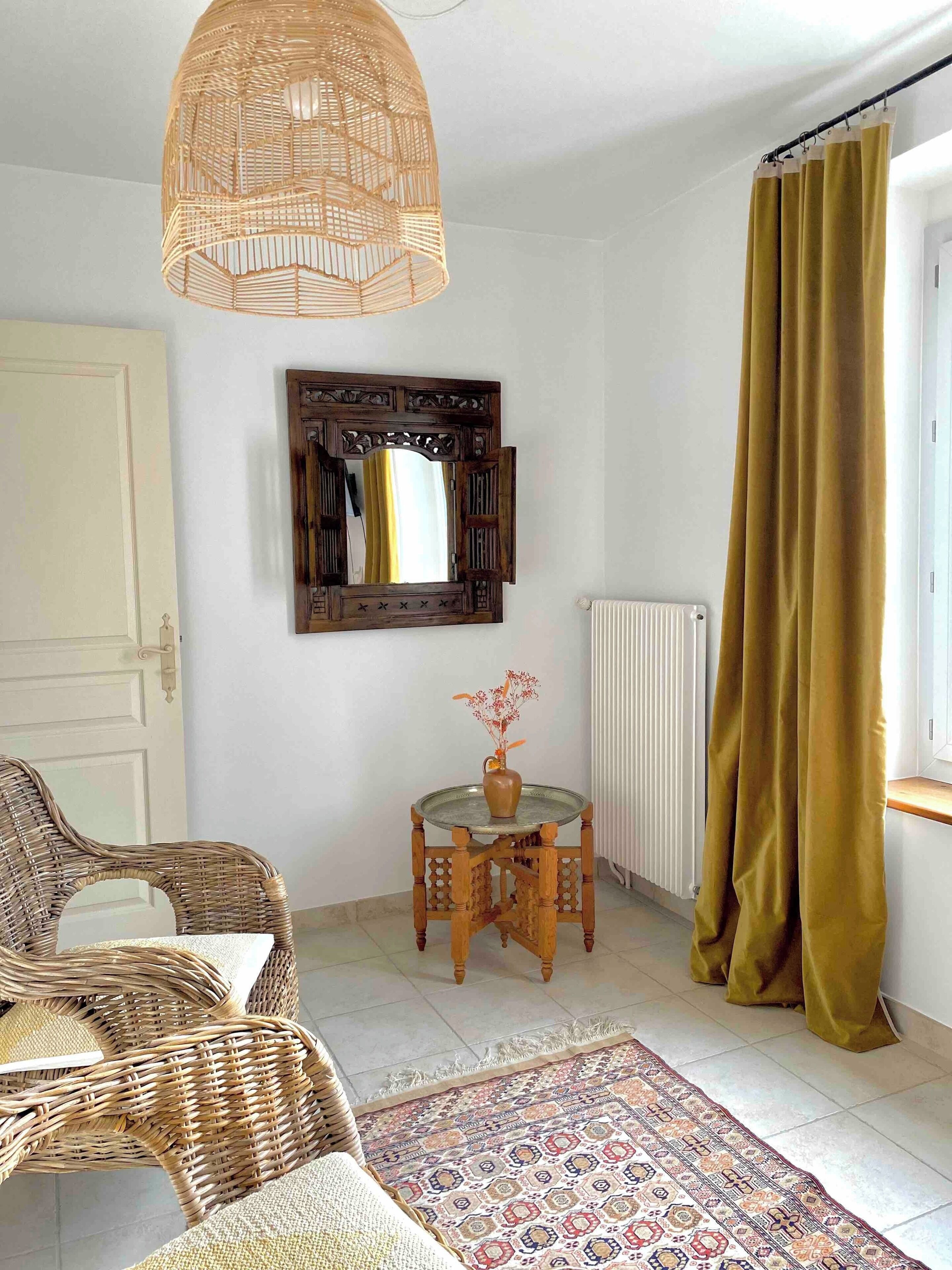 Une pièce comprenant une chaise en osier, une petite table ronde avec un vase, un miroir, un radiateur et un tapis à motifs.