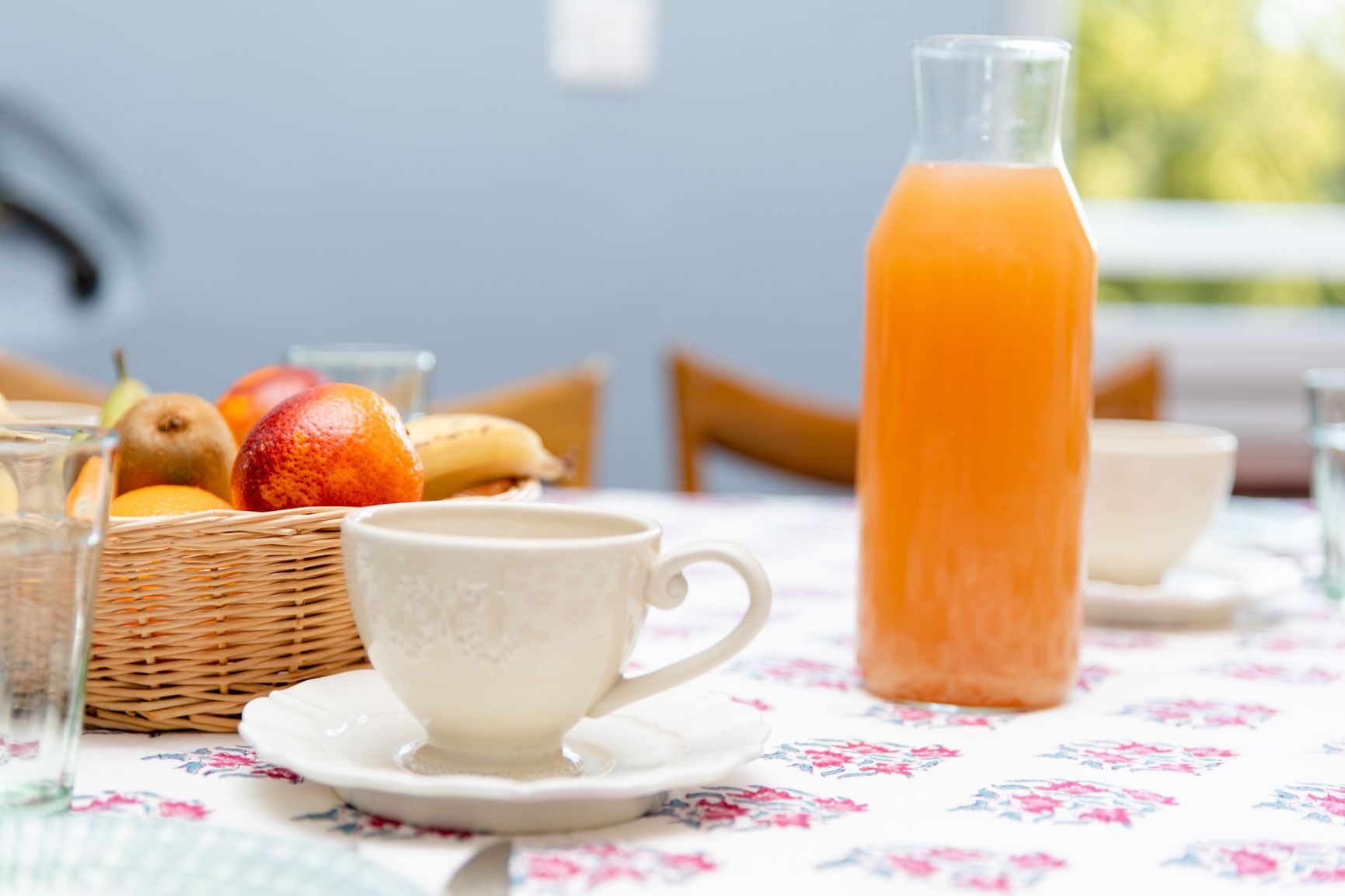 A breakfast setting with a wicker basket of fruits, a white cup, saucer, and a glass of orange juice on a floral tablecloth.