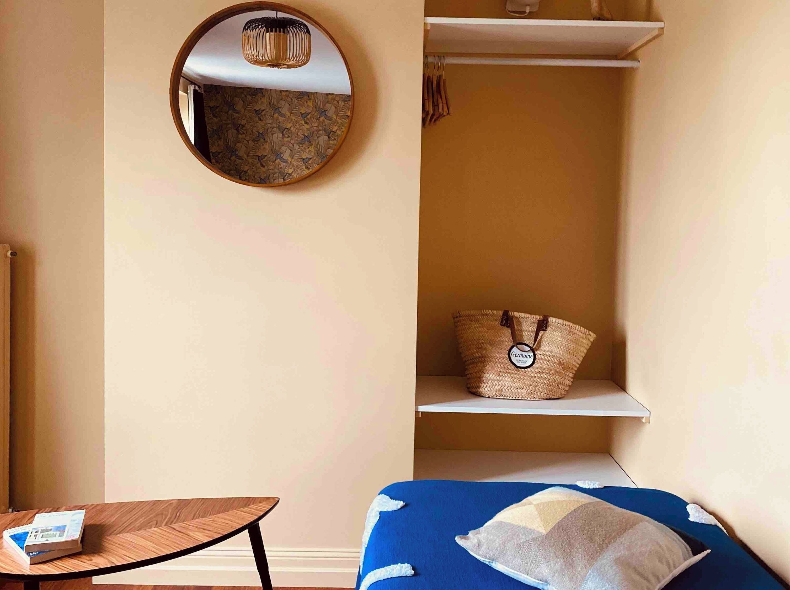 A compact room with a round mirror, a small table, a bed with a blue cover, and a wicker basket on a shelf.