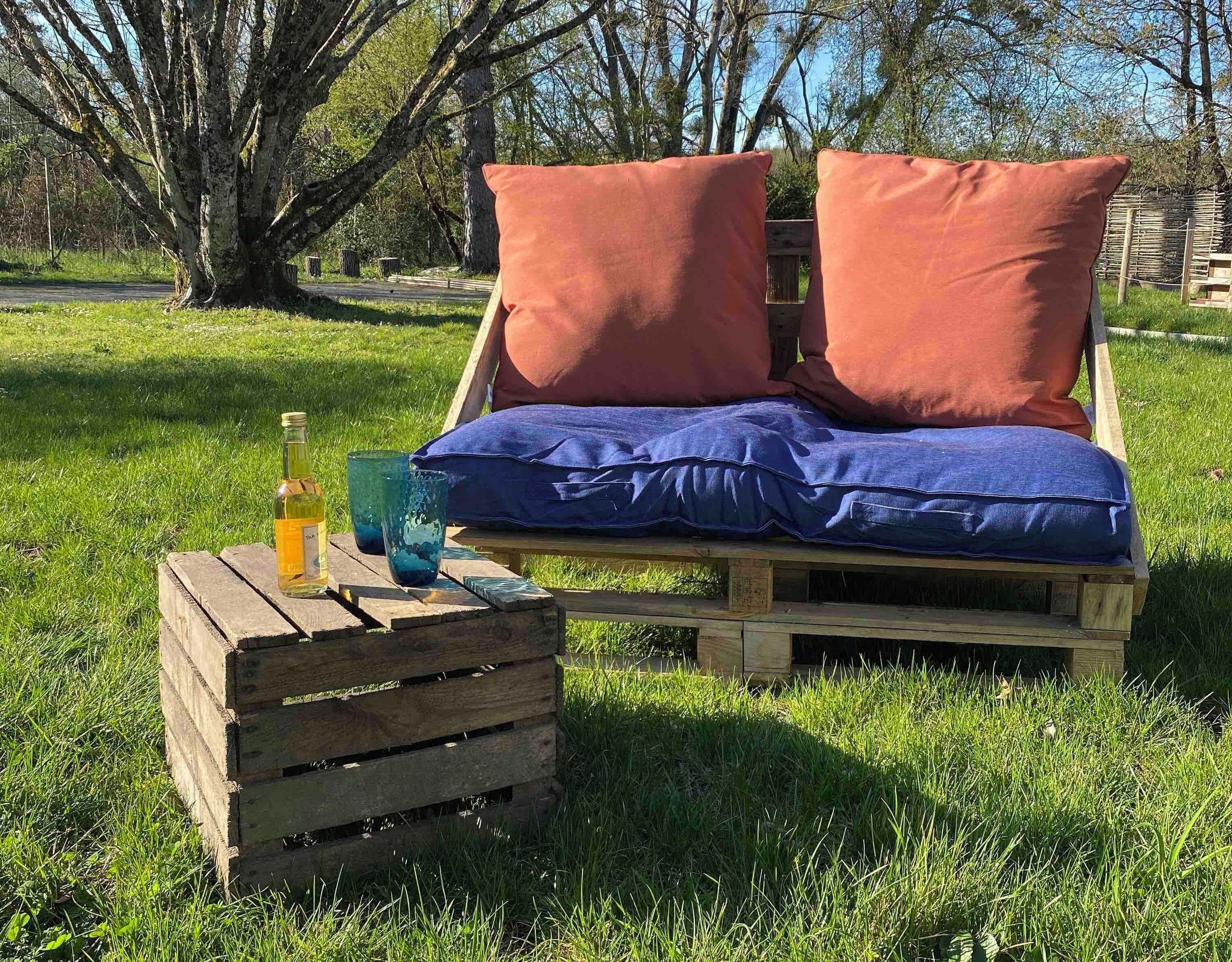 A wooden bench with two orange cushions and a blue cushion, placed on a grassy area with a bottle and a glass on a wooden crate beside it.