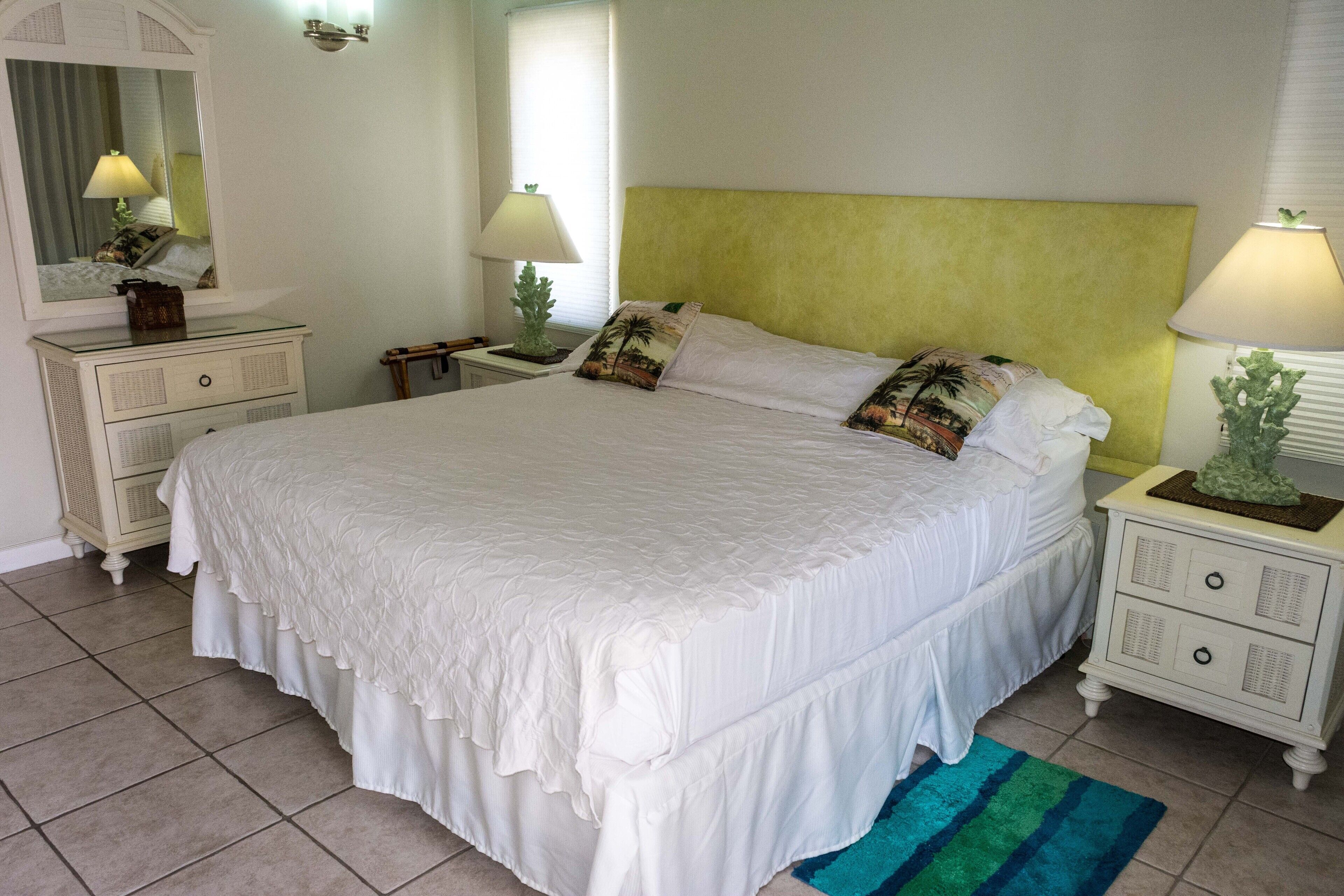 A large bed with a white bedspread and multiple pillows, flanked by two bedside tables with lamps, a dresser, and a mirror.