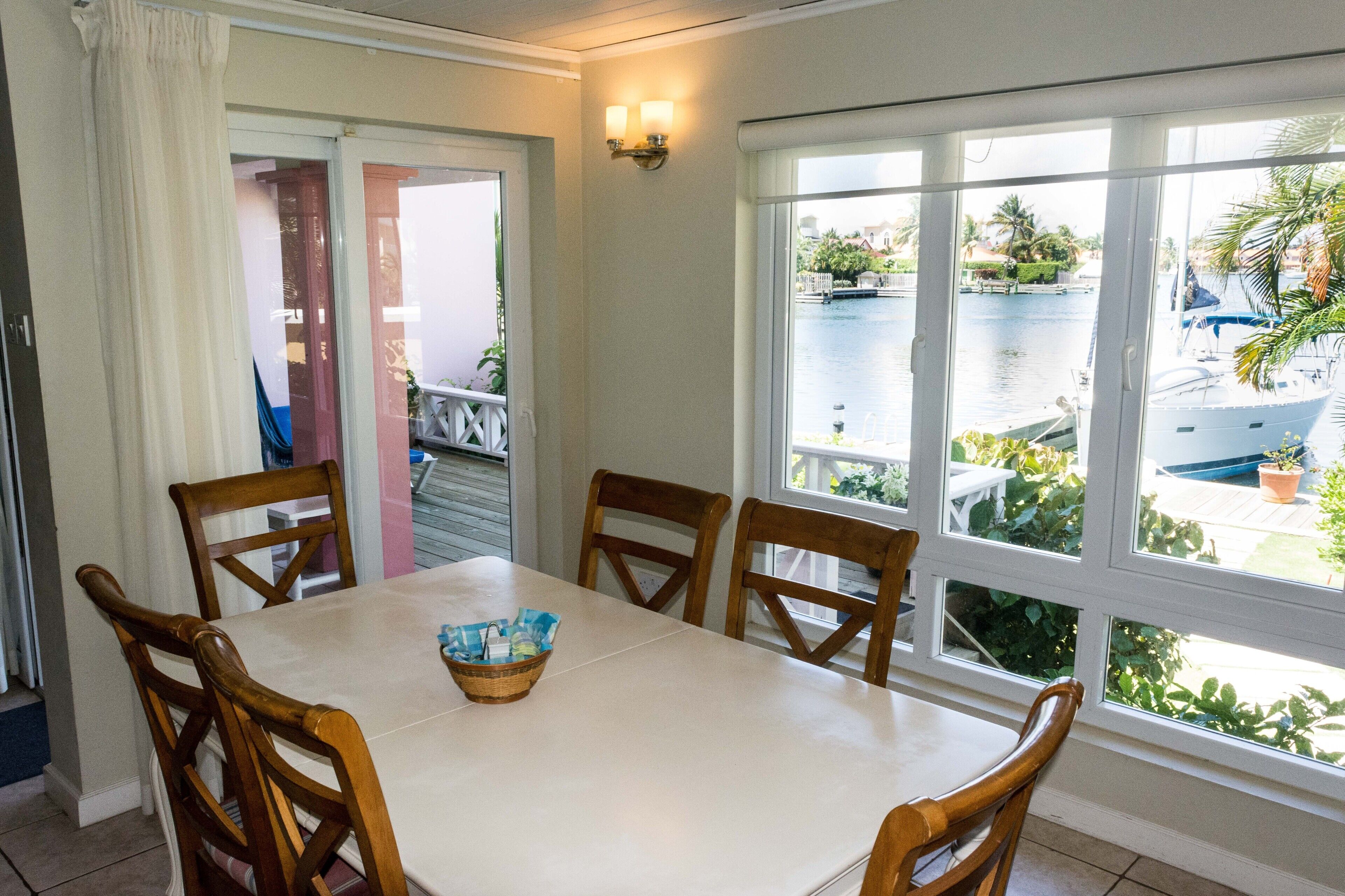 A dining area with a white table and wooden chairs, a basket on the table, and a view of a water body and boats outside.
