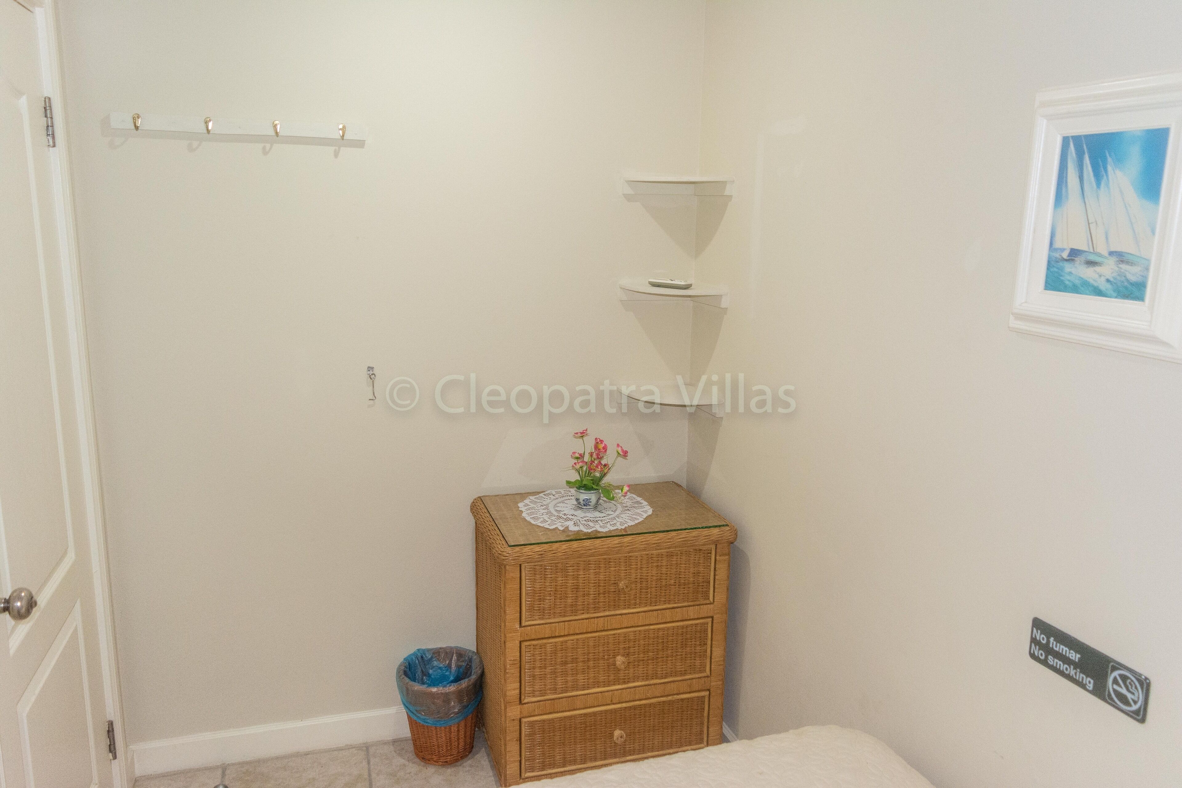A small, clean room with a wicker dresser, a small shelf, a trash bin, and a "No smoking" sign.