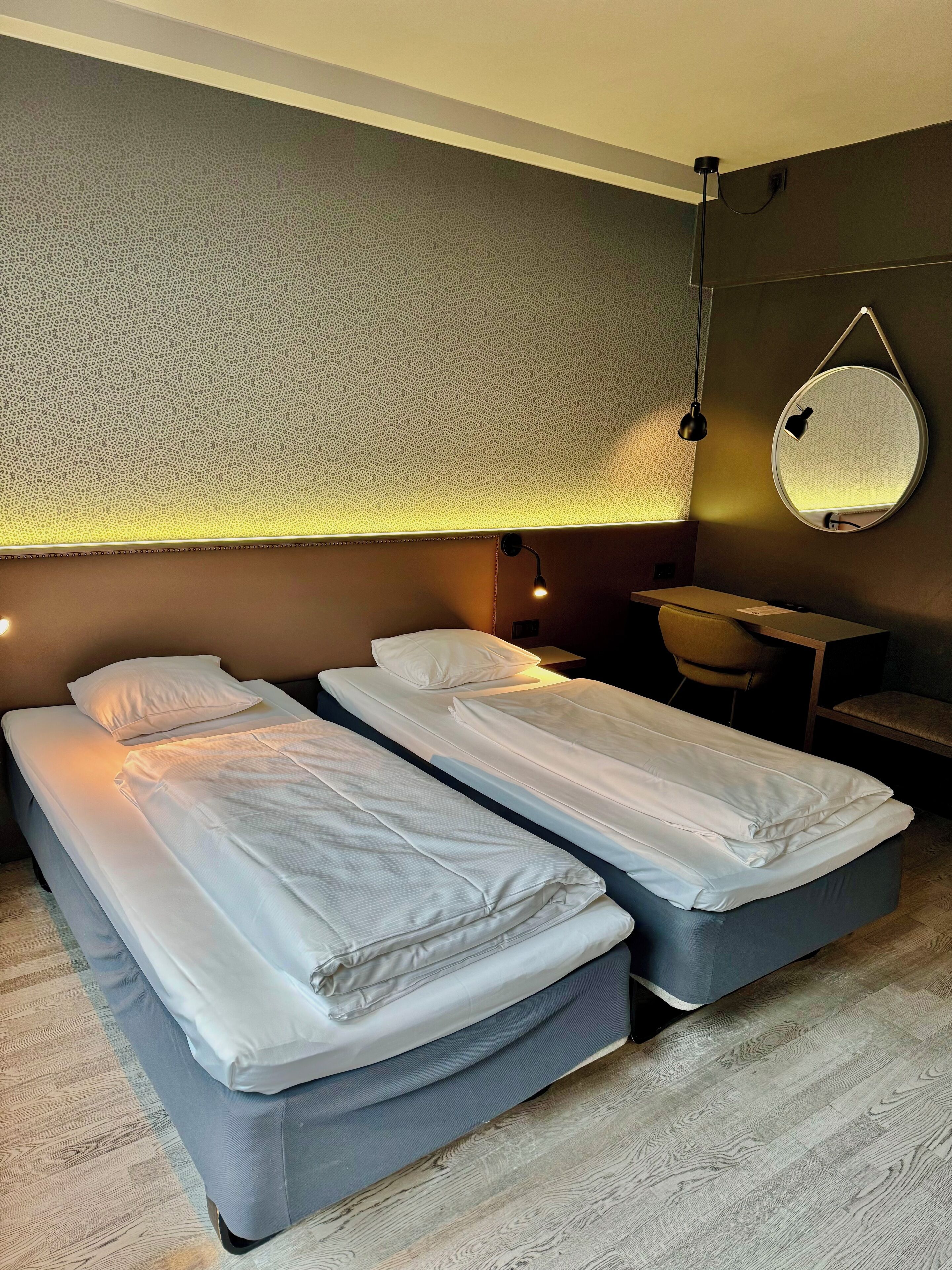 Et hotelværelse med to enkeltsenge, et skrivebord, en stol og et rundt spejl på væggen.