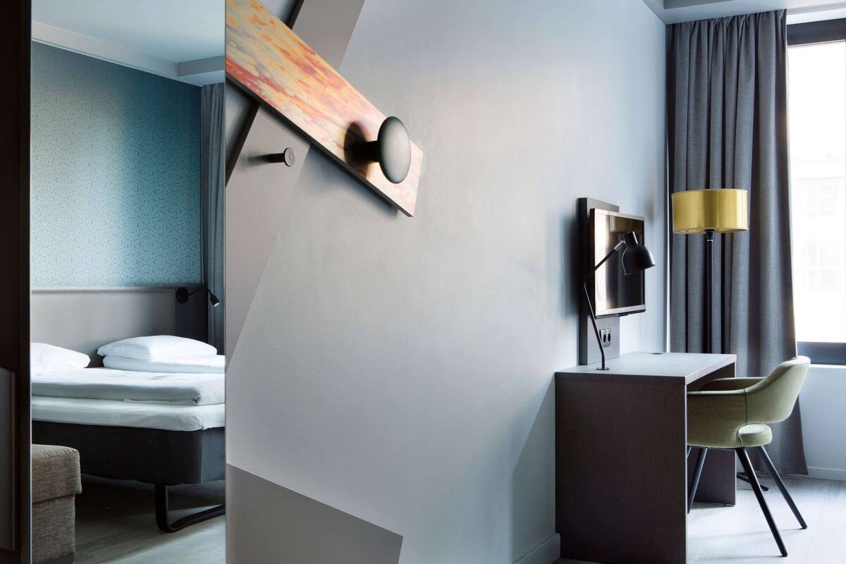 Moderni hotellihuone, jossa on sänky, työpöytä, tuoli ja suuri ikkuna, jossa on verhot.