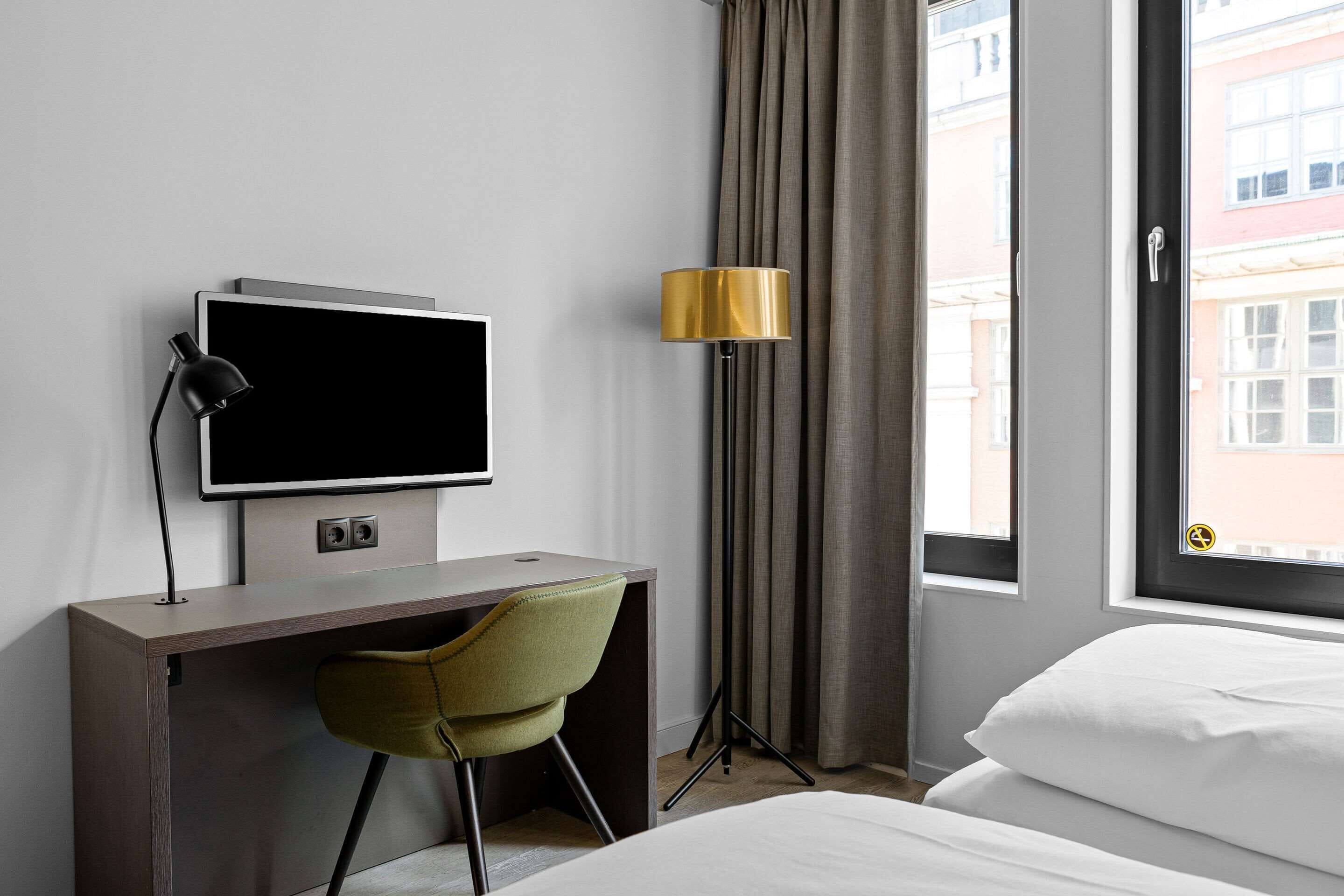 Habitación de hotel con cama, escritorio con televisor, una silla, lámpara y ventana con cortinas.