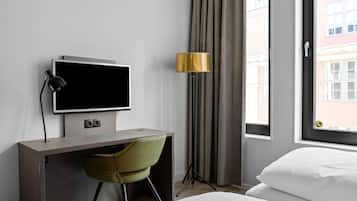 Habitación de hotel con cama, escritorio con televisor, una silla, lámpara y ventana con cortinas.