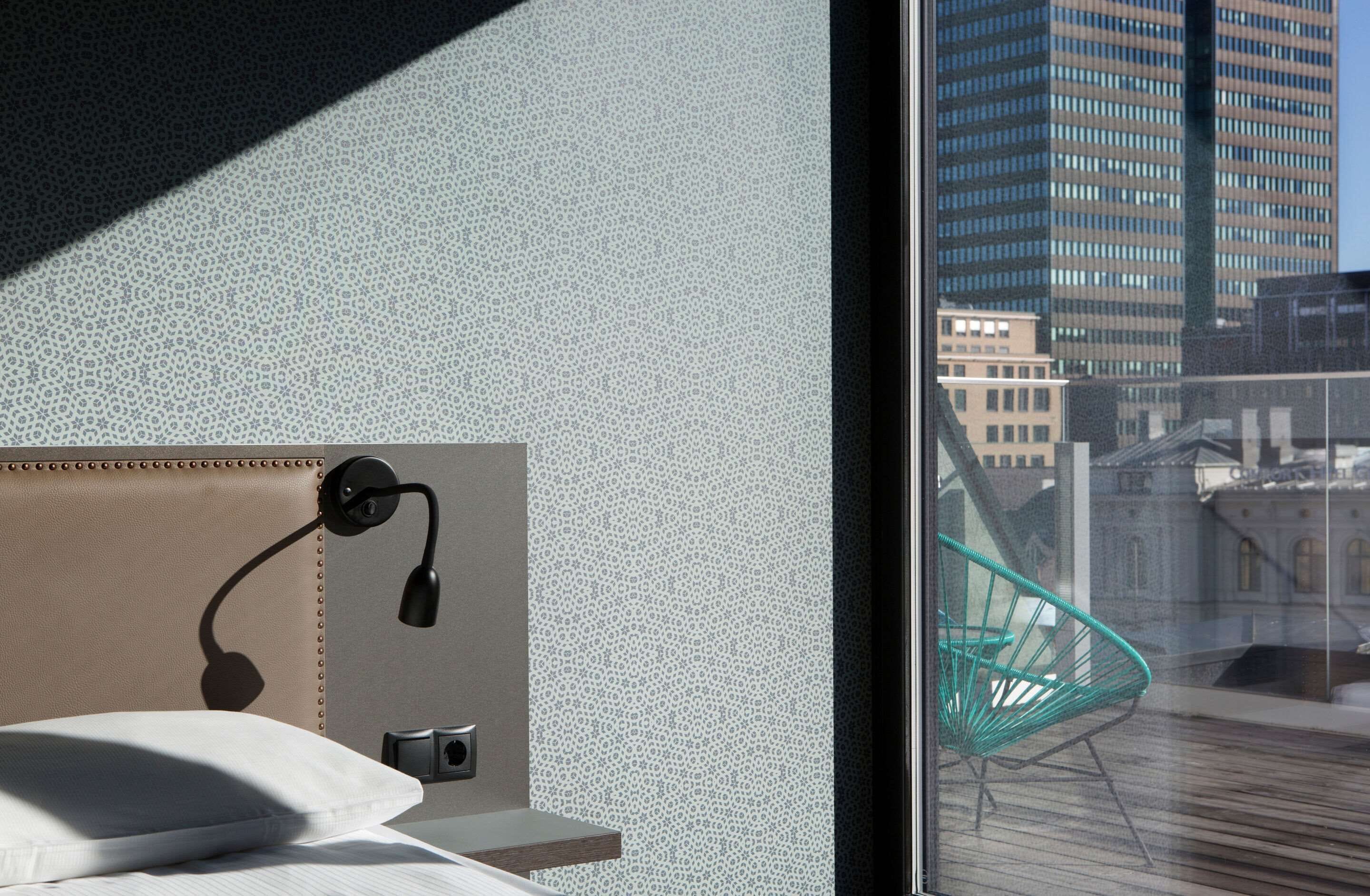 A hotel room with a patterned wall, a bed with a pillow, a desk lamp, and a view of a cityscape through a window.