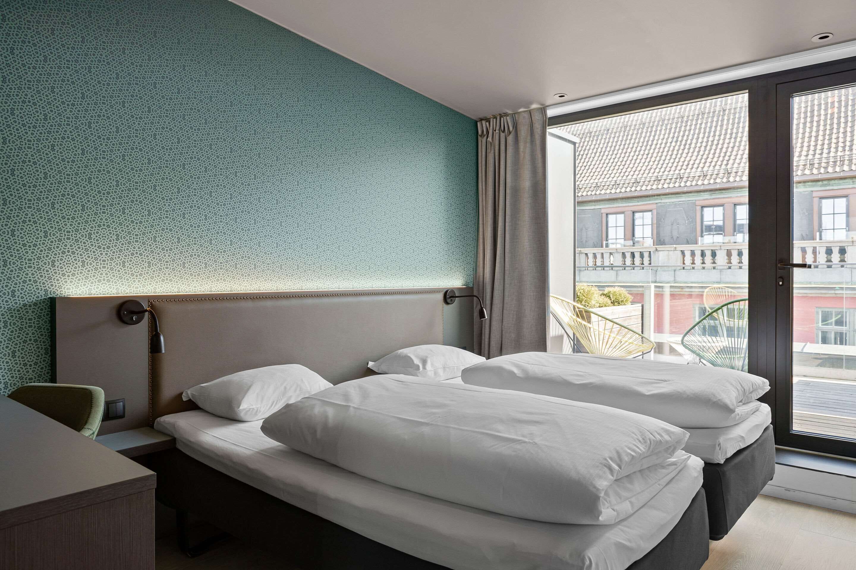 A hotel room with a large bed, a desk, and a view of the city through a sliding glass door.