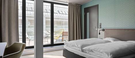 A modern hotel room with a large bed, a desk, and a view of the outdoor terrace.