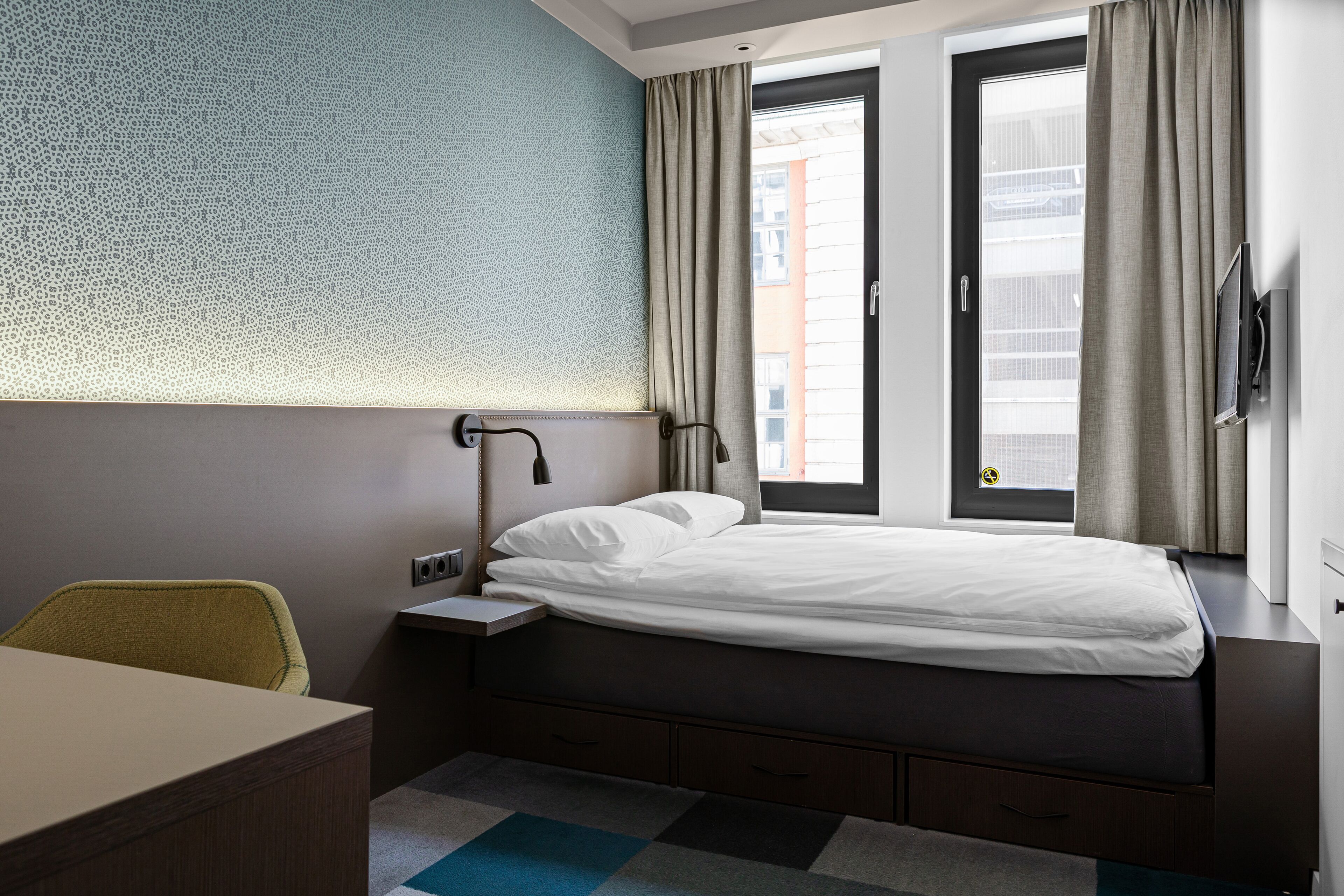 A modern hotel room with a bed, a desk, and a chair. There is a window with curtains, a wall-mounted TV, and a patterned wall.