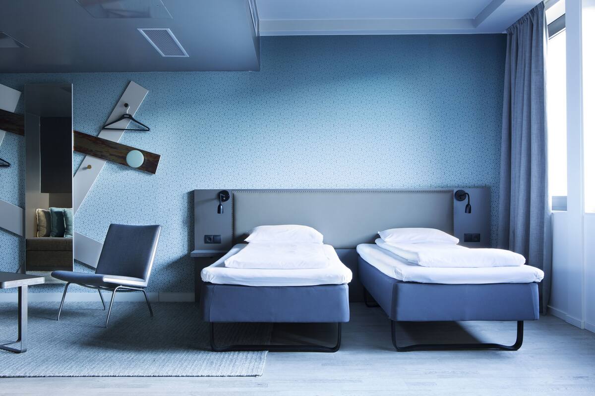 Moderni hotellihuone, jossa on kaksi erillistä sänkyä, työpöytä, tuoli ja suuri ikkuna.