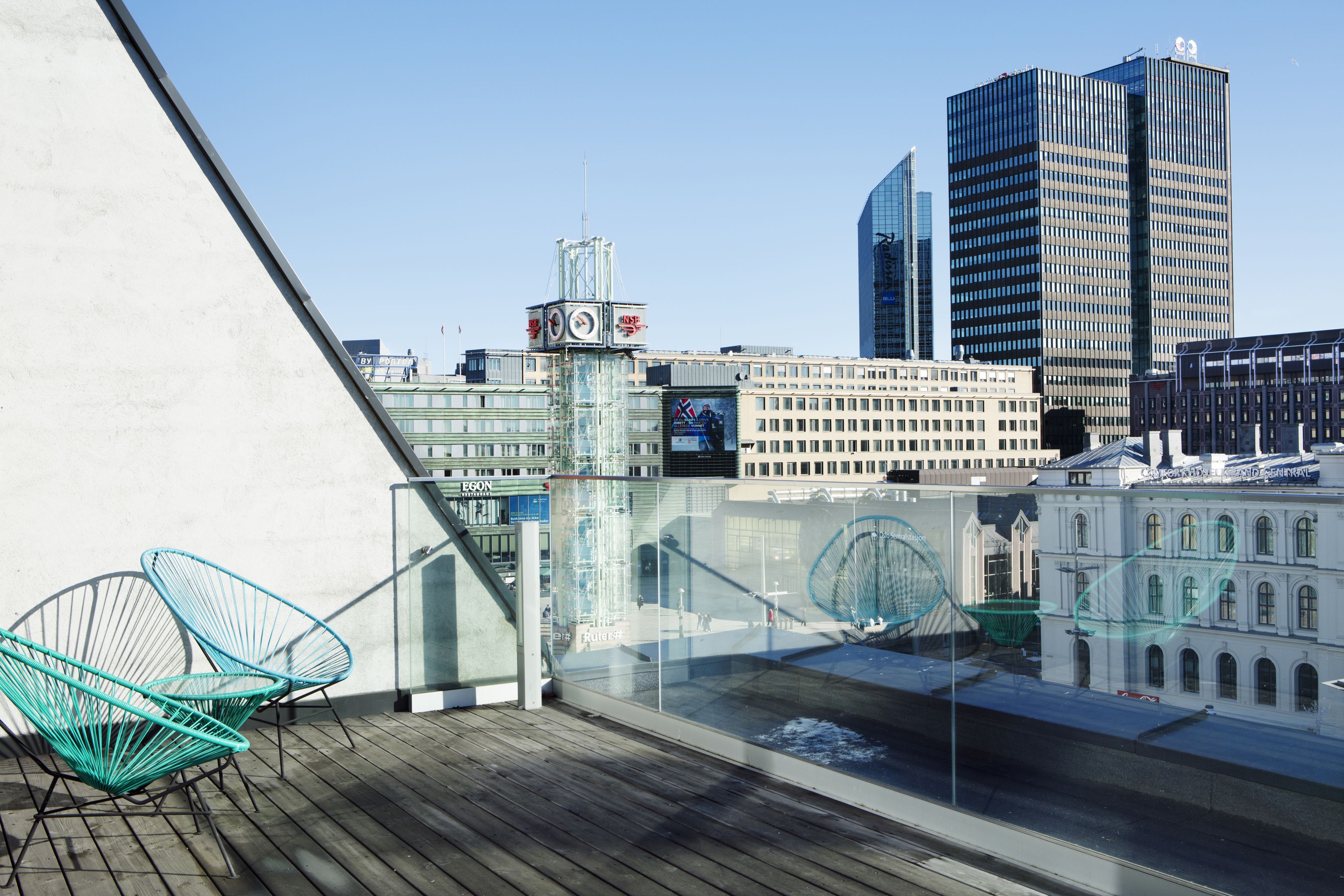 A city view from a balcony with modern chairs, a glass railing, and a view of historic and modern buildings.