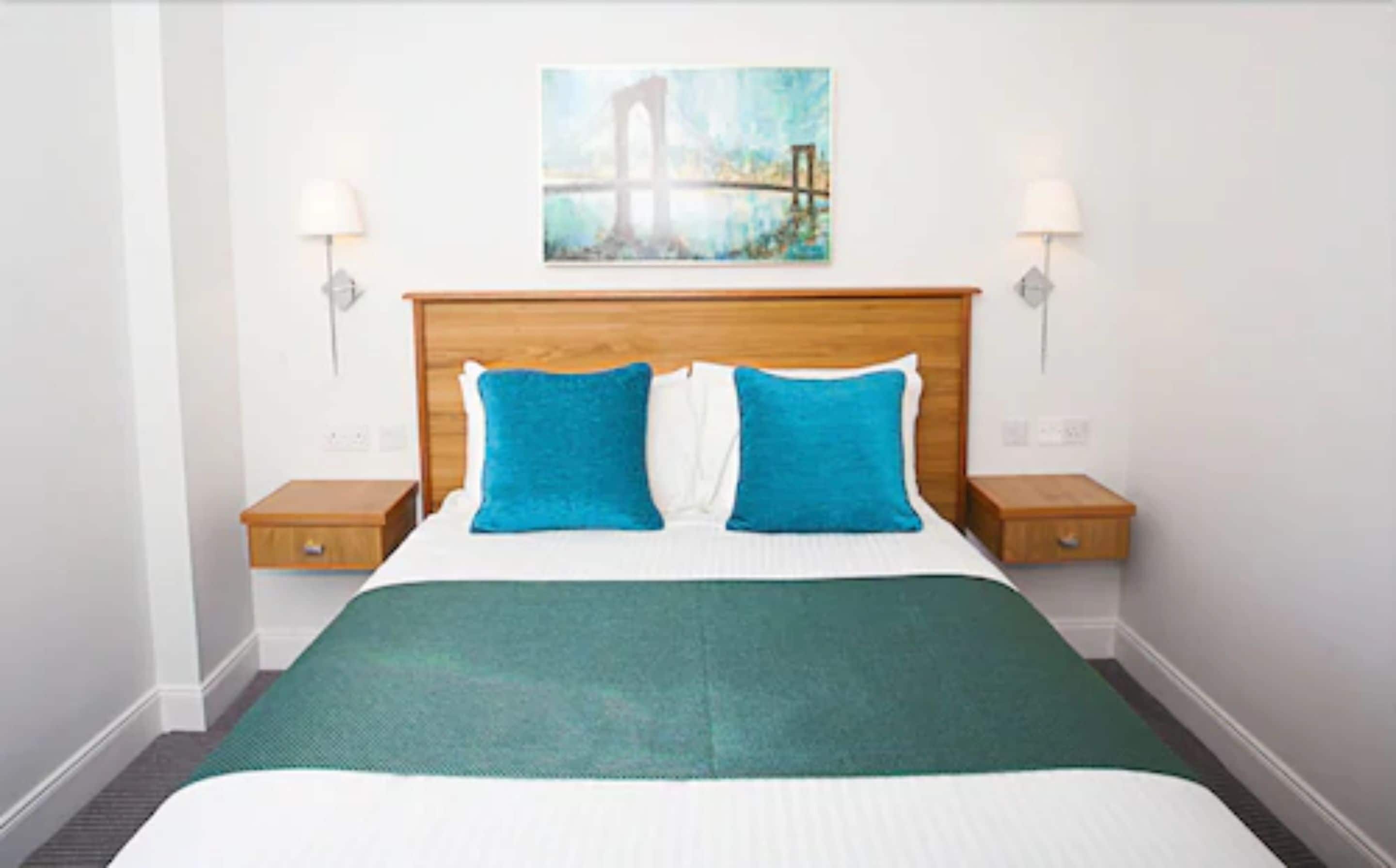 A hotel room with a wooden headboard, two blue pillows, a green bedspread, and a painting of a bridge on the wall.