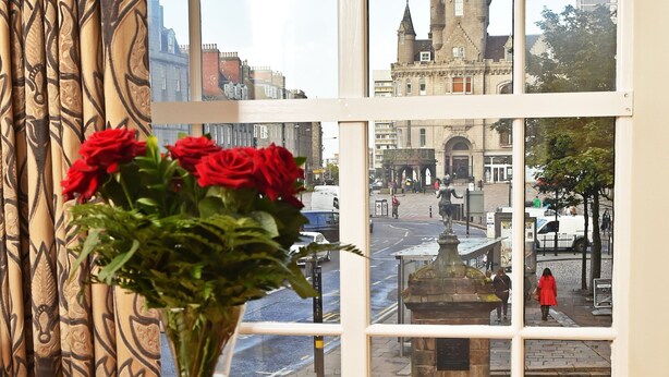 A room with a large window, a vase with red roses, and a book on a glass table. - Royal Athenaeum Suites (Aberdeen)