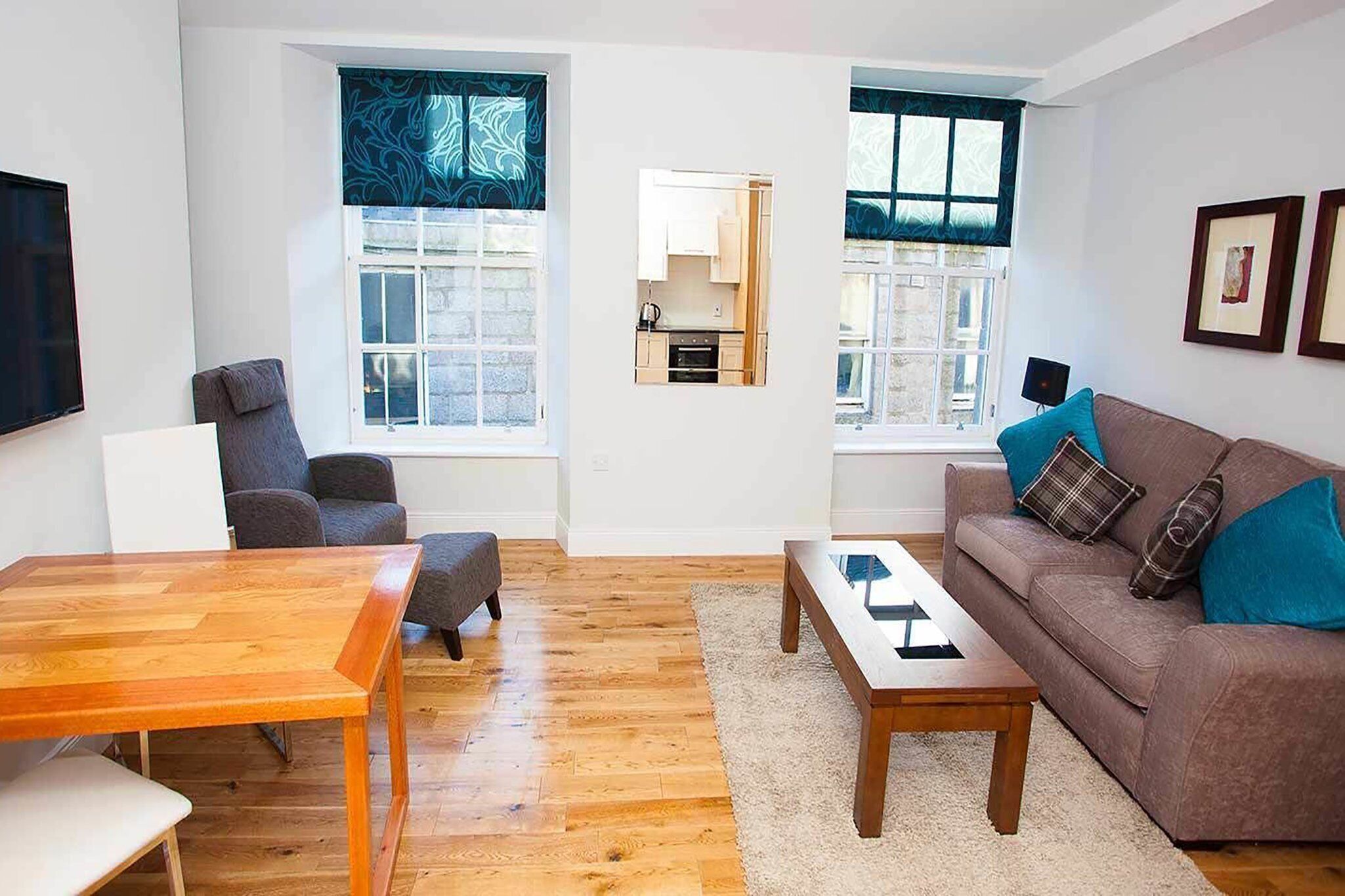 A modern living room with a wooden dining table, a sofa with cushions, and a view of the kitchen through a window.