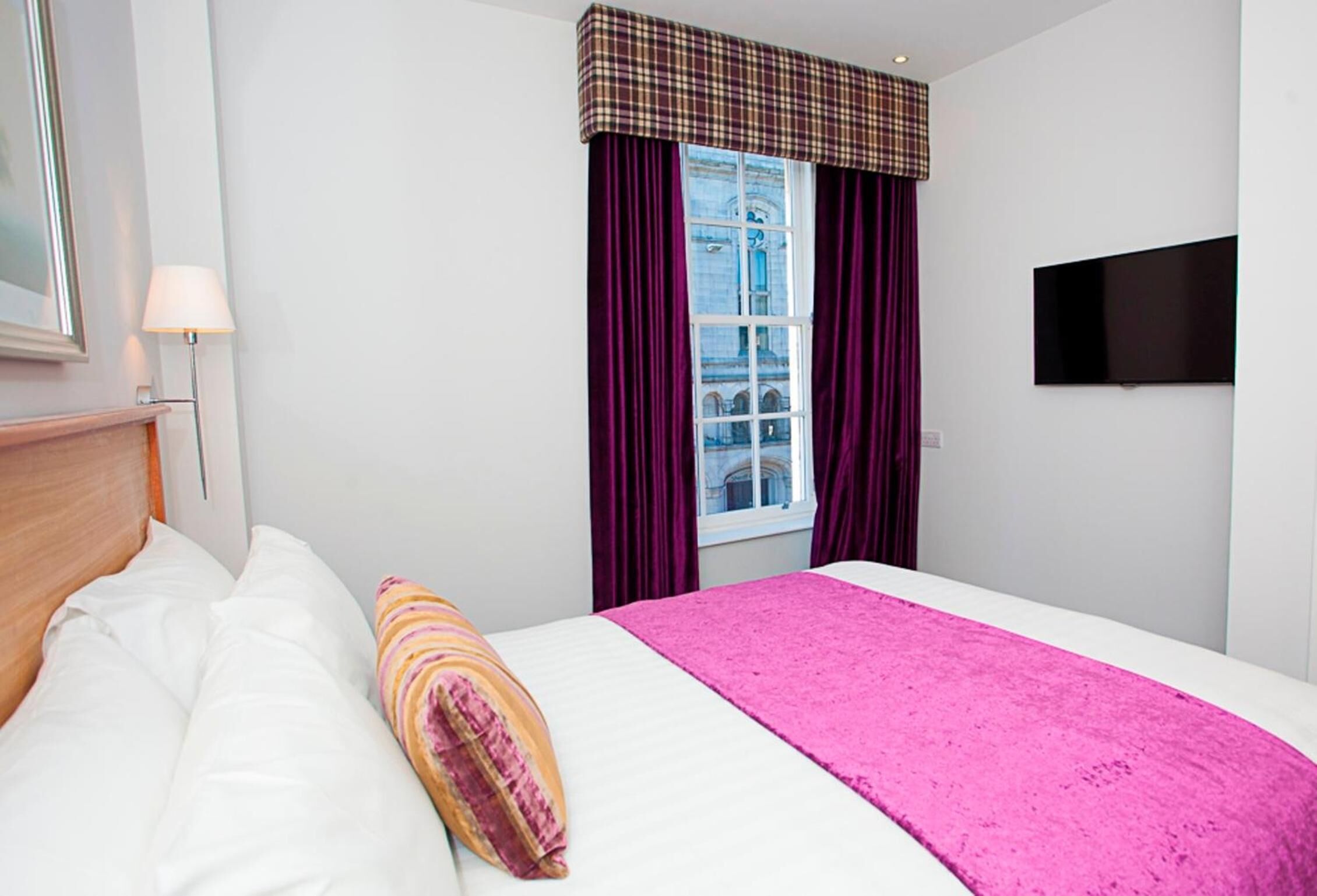 A hotel room with a bed, a window with purple curtains, a television mounted on the wall, and a lamp beside the bed.