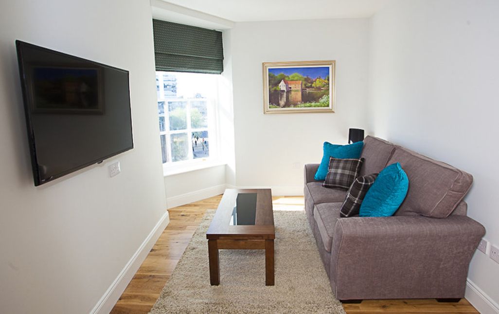 Uma sala de estar com uma televisão de tela plana, um sofá cinza com almofadas azuis, uma mesa de centro de madeira e um quadro pendurado na parede.
