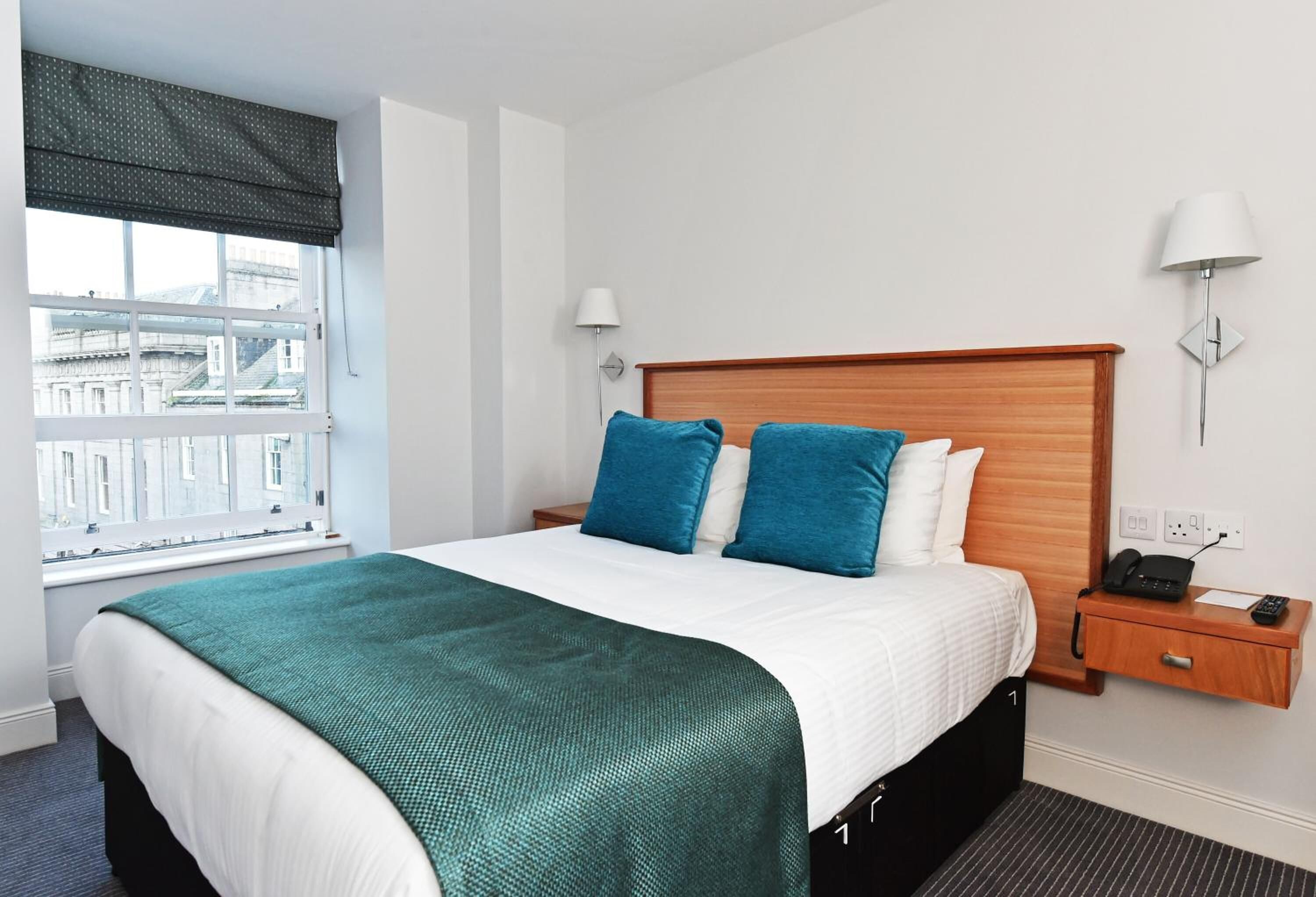 A hotel room with a large bed, a window with a view of buildings, a bedside table with a lamp, and a phone.