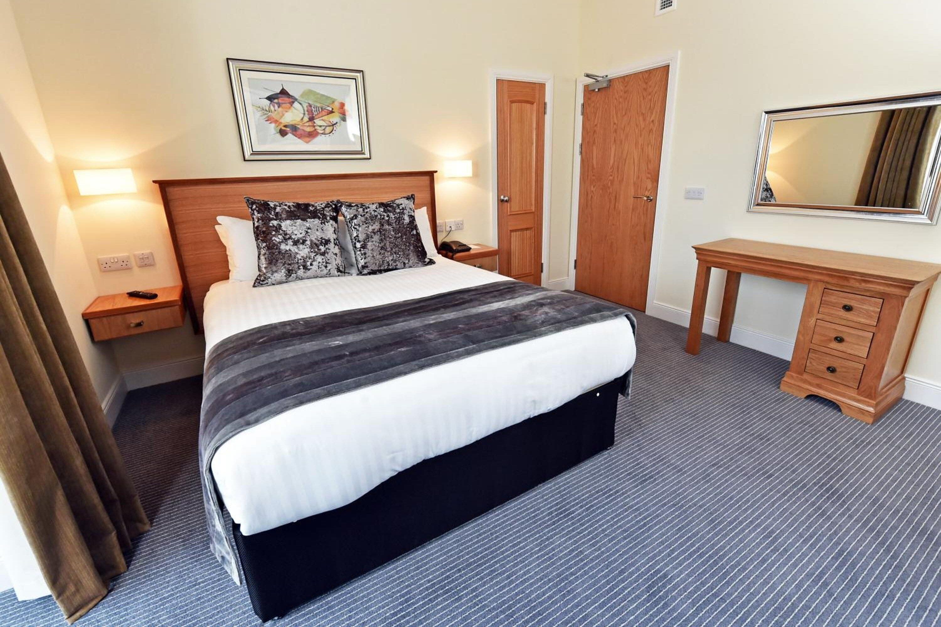Habitación de hotel con una cama grande, un aparador de madera, una mesita de noche, un cuadro enmarcado en la pared y una puerta.