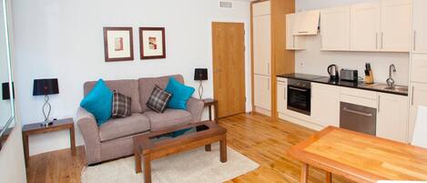 A modern living room with a sofa, coffee table, and a dining table. There is a kitchen area with cabinets and appliances.