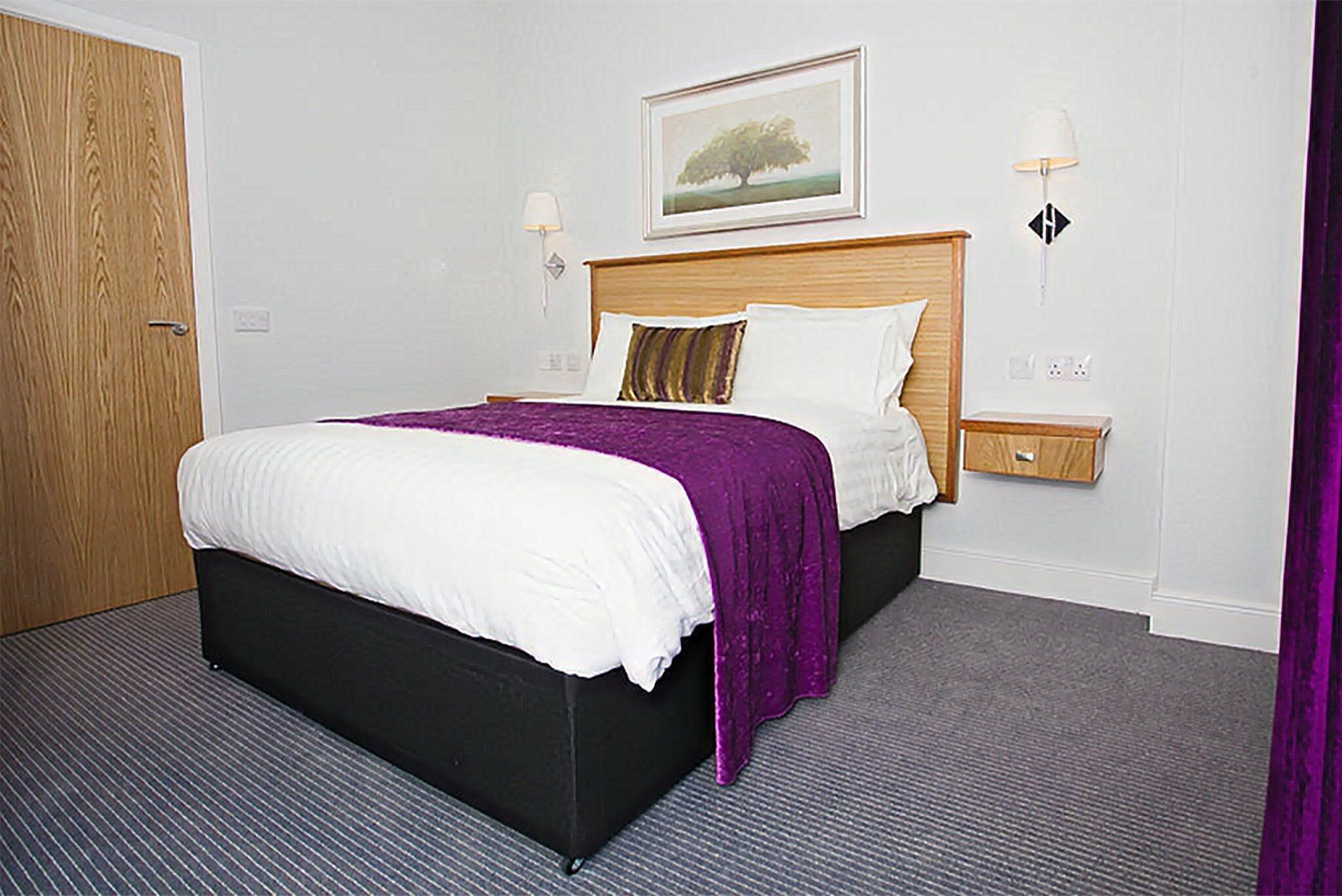 A hotel room with a large bed, a wooden headboard, a nightstand, and a framed picture on the wall.