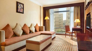 A hotel room with a sofa, a coffee table, a TV, a desk with a lamp, and a window with curtains. - Ramada by Wyndham Riyadh (Riyadh)