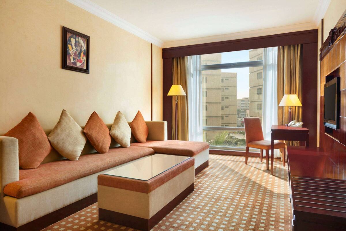 Habitación de hotel con sofá, mesa de centro, televisor, escritorio con lámpara y ventana con cortinas.