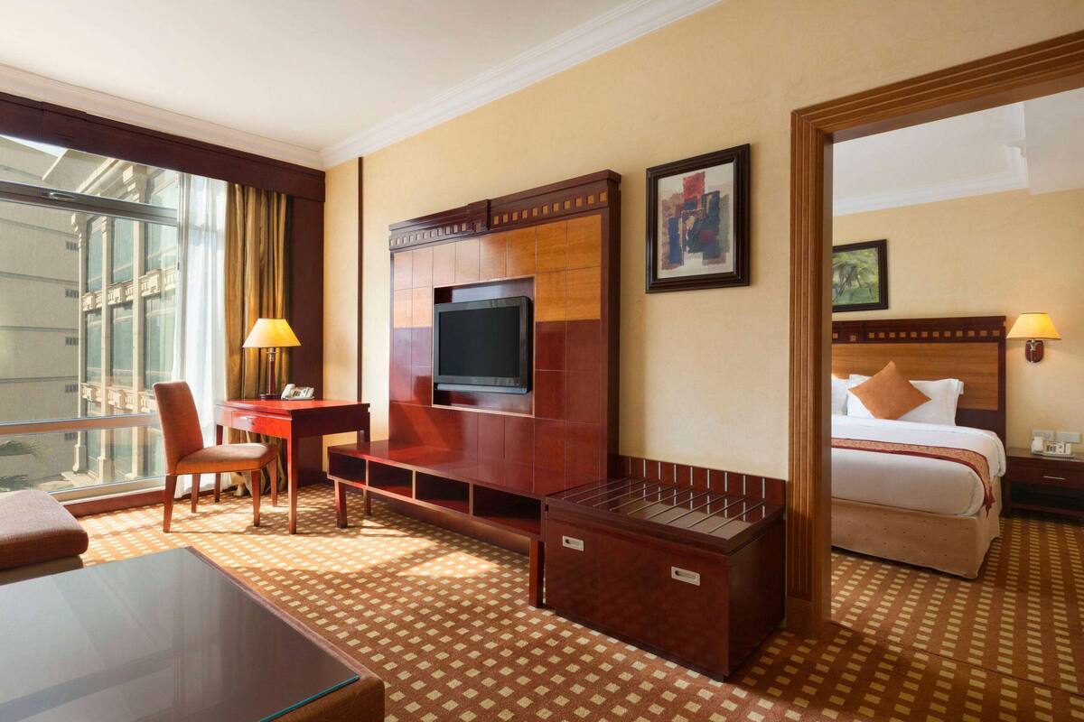 Habitación de hotel con una televisión grande, un escritorio, una silla, una cama y vista a la ciudad por la ventana.