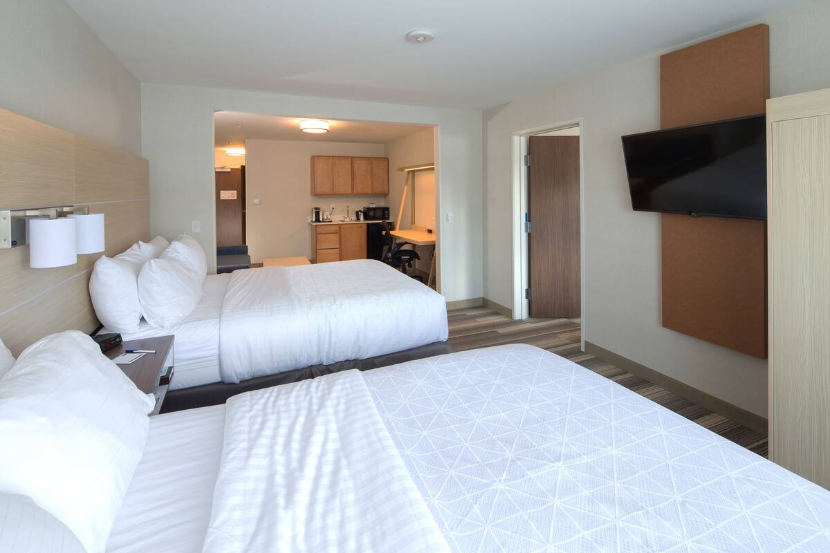 Habitación de hotel con dos camas, televisión, escritorio y una kitchenette al fondo.