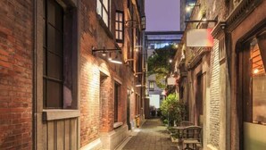 Une ruelle étroite bordée d’immeubles en briques, avec des tables et des chaises installées à l’extérieur et des guirlandes lumineuses suspendues.