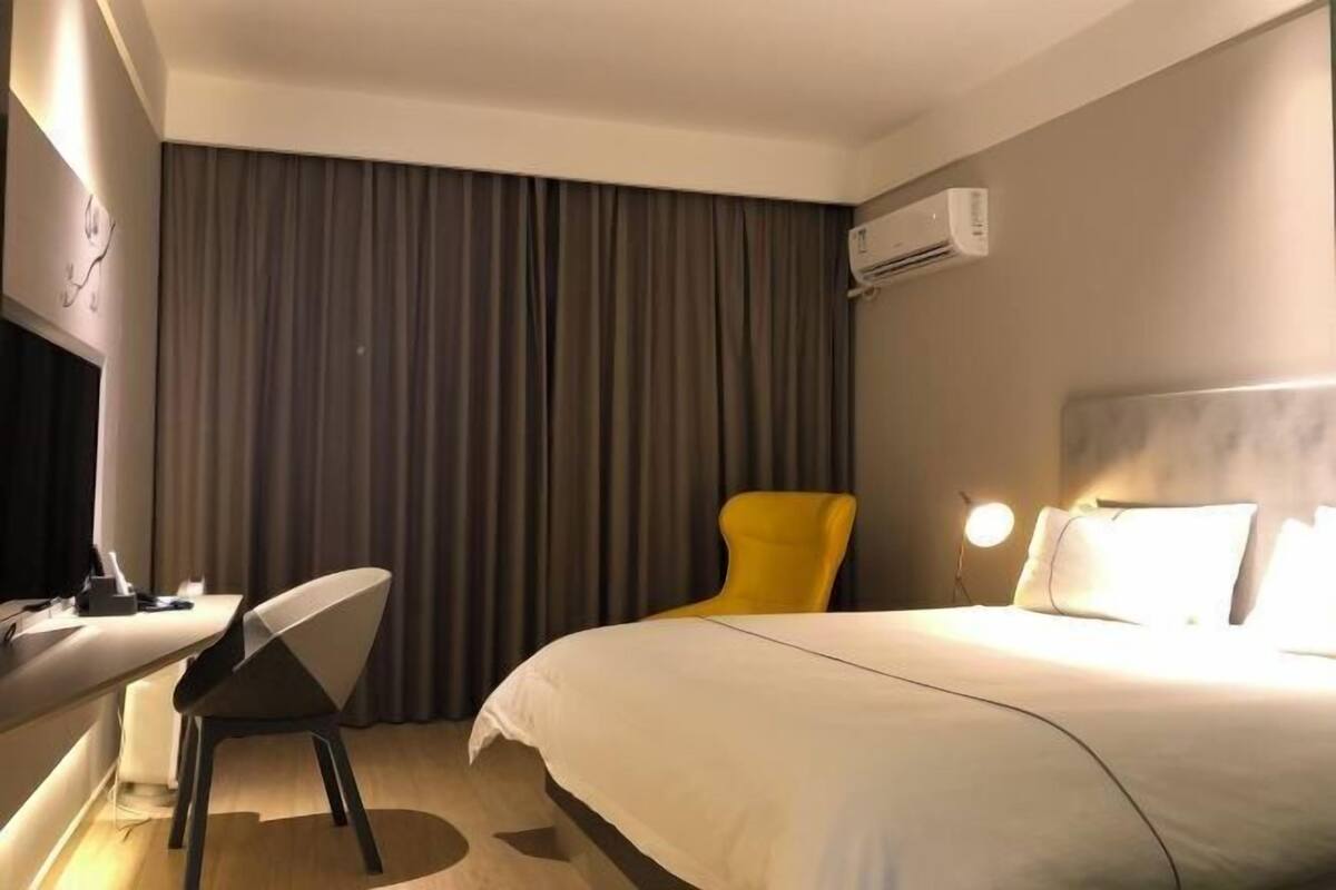A hotel room with a bed, a desk with a chair, a yellow chair, a lamp, and an air conditioning unit.