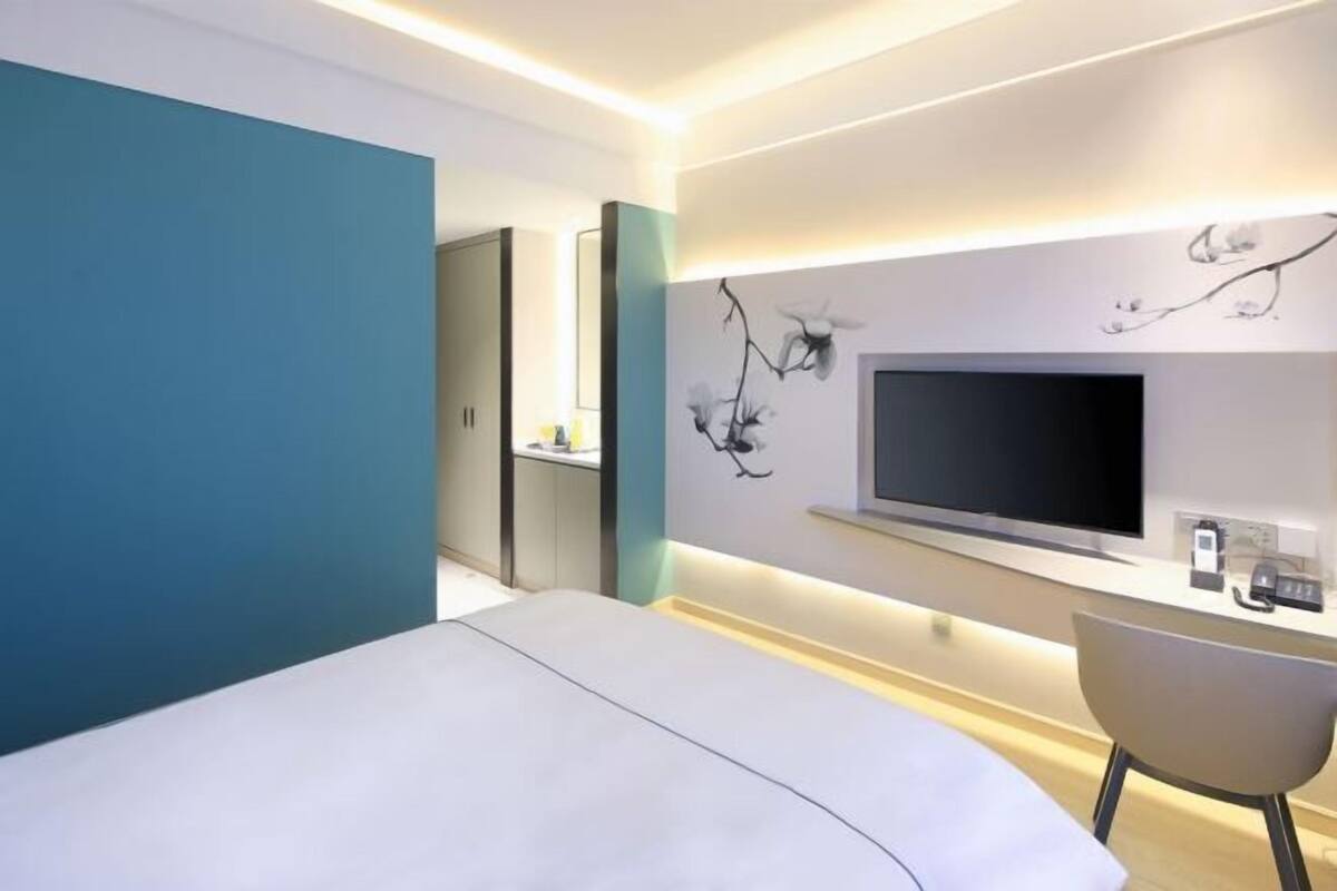 A modern hotel room with a bed, a desk with a flat-screen TV, a chair, and a wall-mounted artwork.