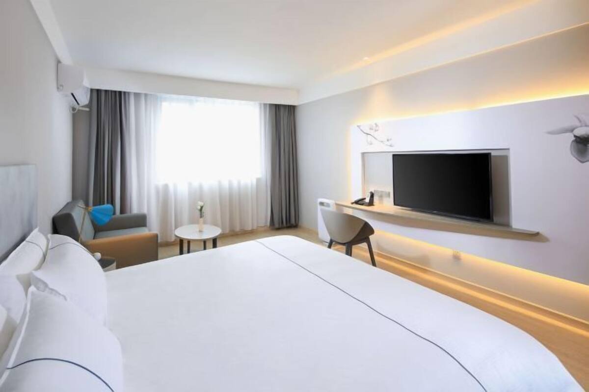 A modern hotel room with a large bed, a flat-screen TV mounted on the wall, a small round table, and a seating area with a sofa and chairs.