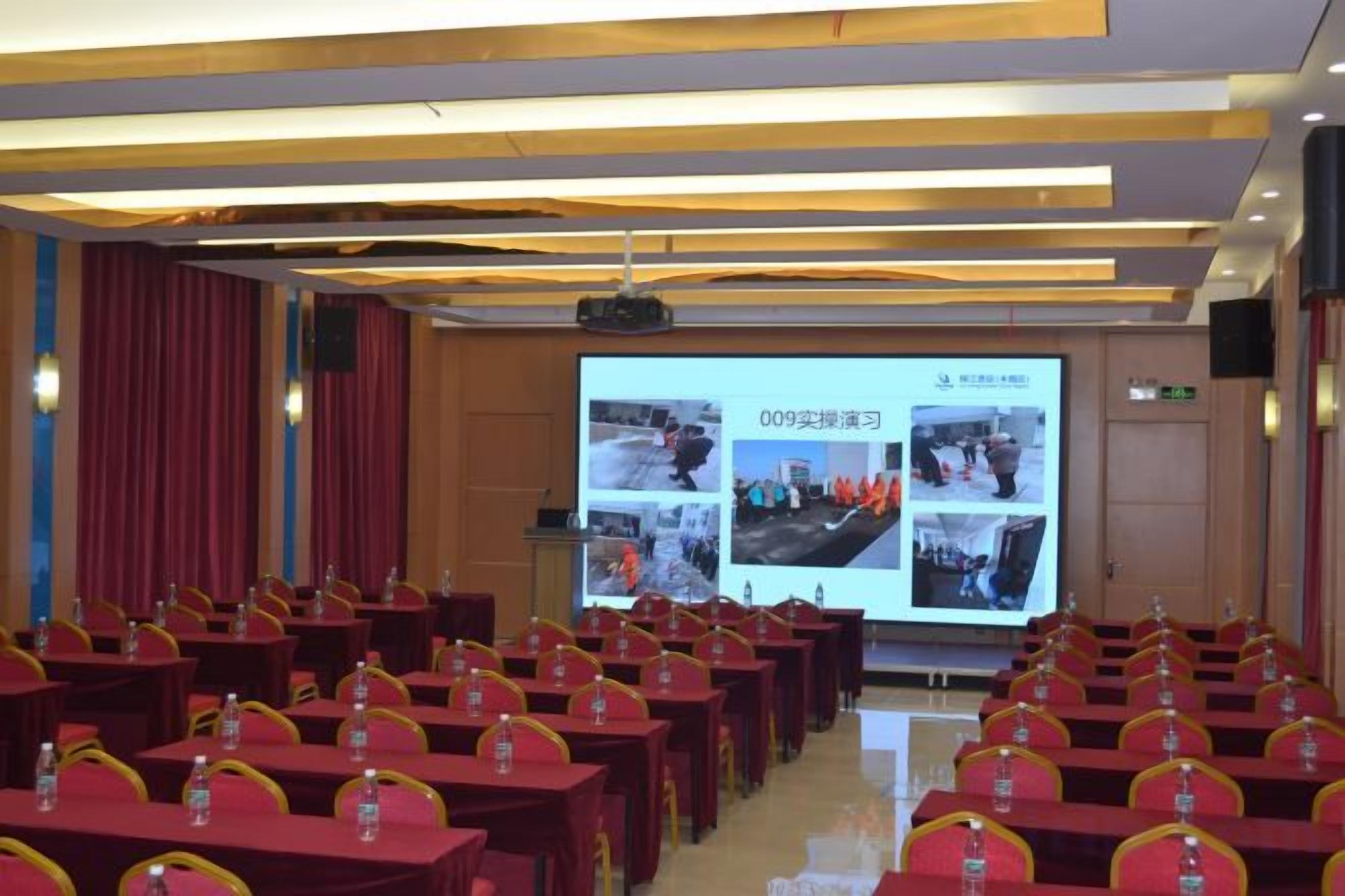 A conference room with rows of tables and chairs, a projection screen displaying images, and a podium at the front.