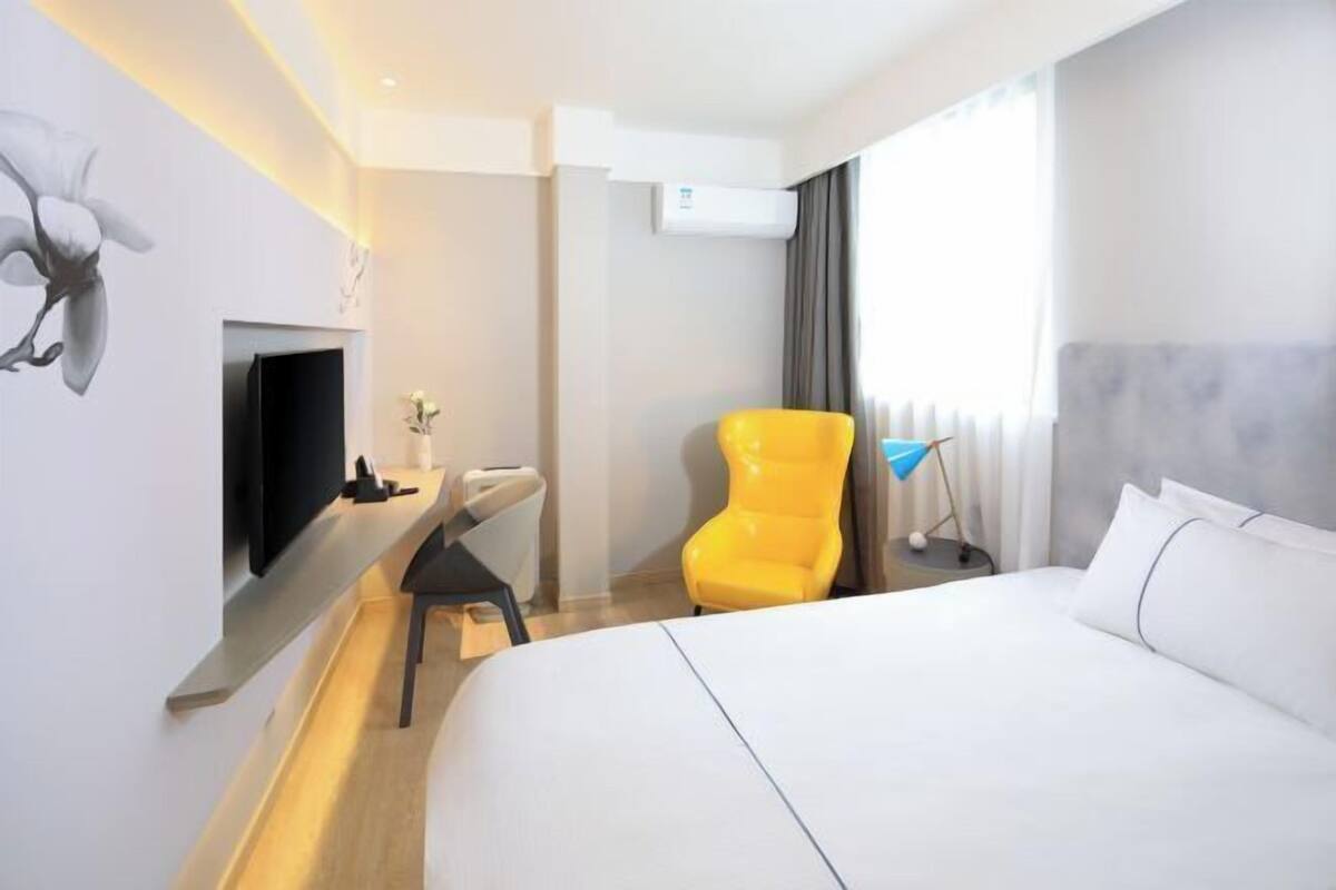 A modern hotel room with a large bed, a flat-screen TV mounted on the wall, a desk with a chair, and a bright yellow armchair.