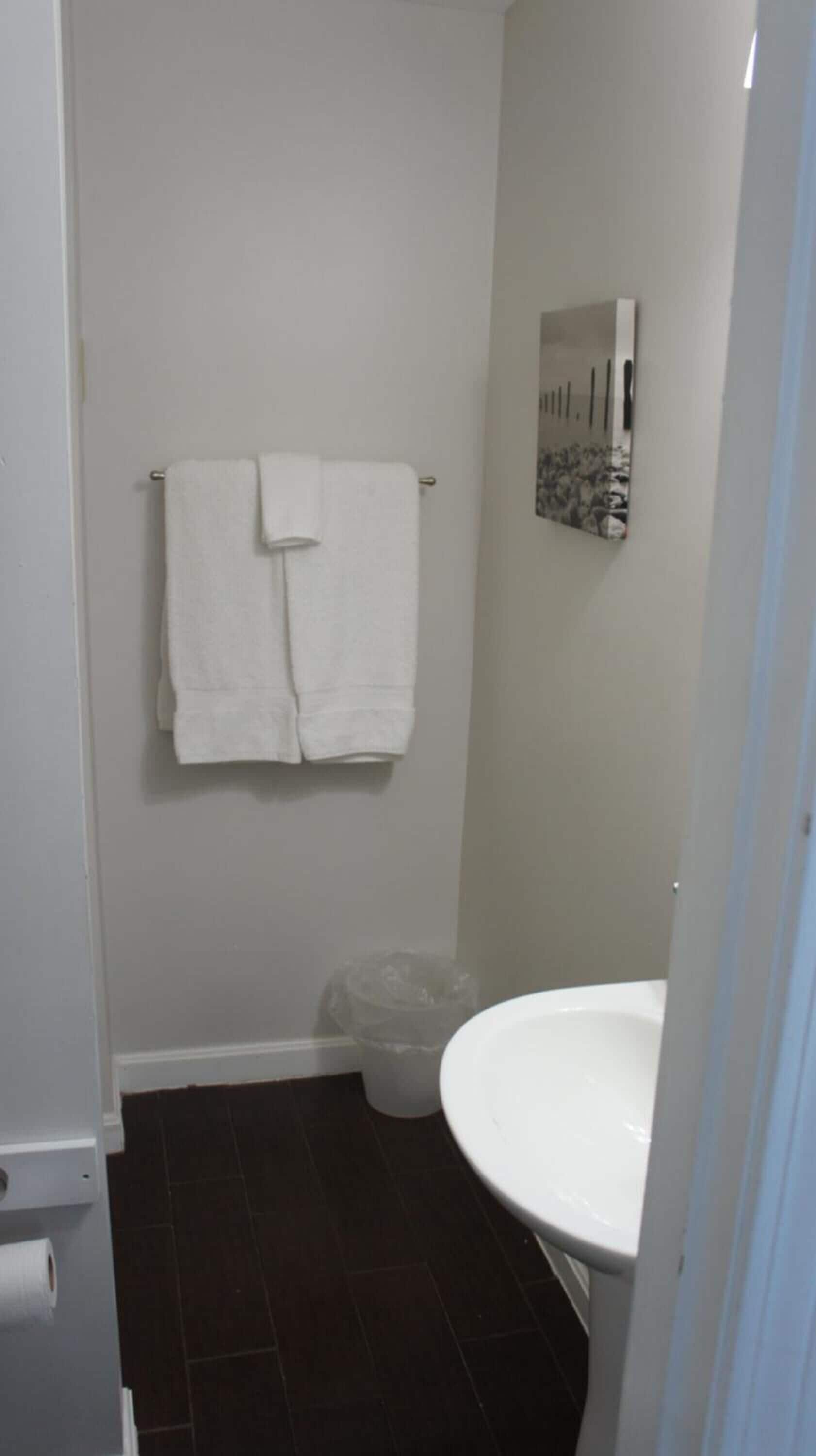 Een kleine badkamer met een wit wastafel, twee witte handdoeken aan een metalen rek, een prullenbak en een spiegel aan de muur.