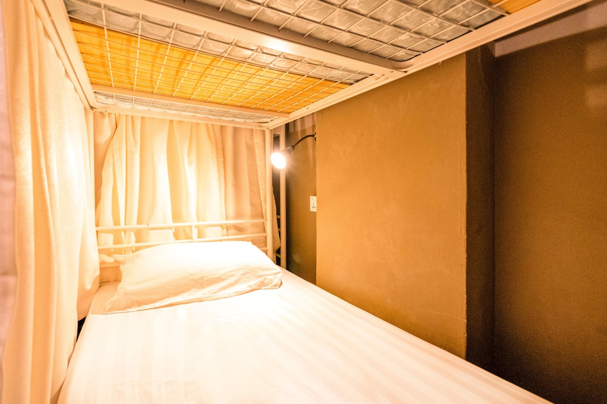 A single bed with a headboard, a window with curtains, and a light fixture on the wall.