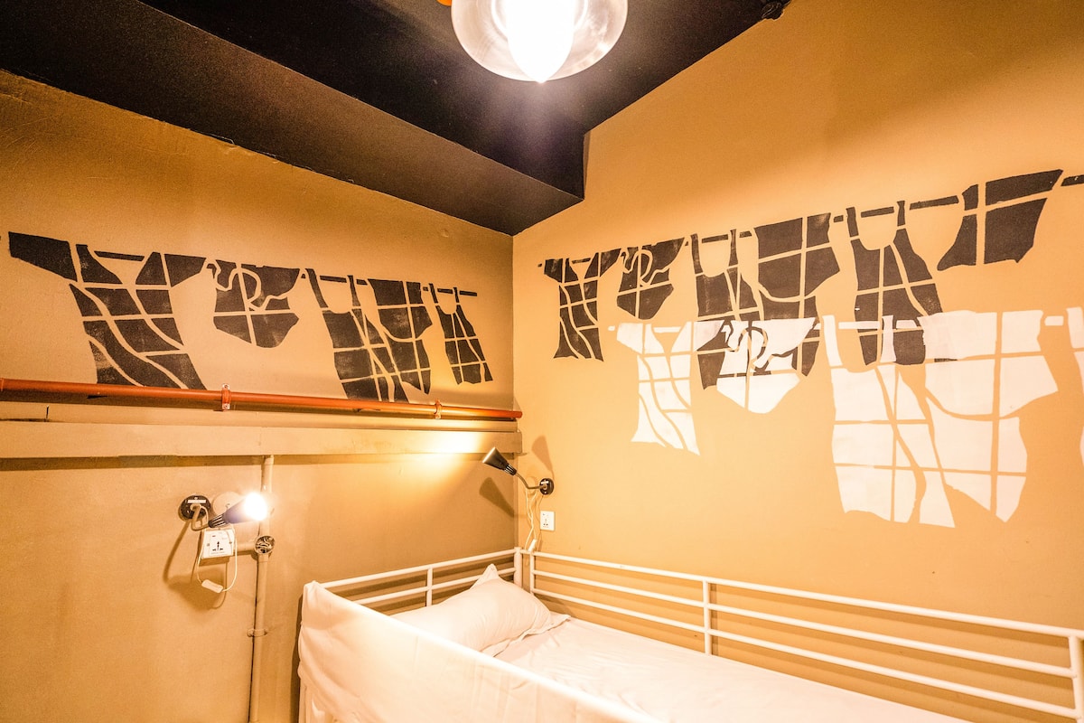 A single bed with a white frame, a wall-mounted lamp, and a power outlet.
