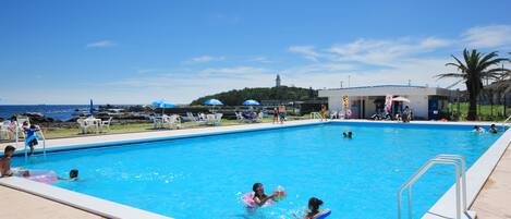 Una piscina con gente nadando y jugando, rodeada de sillas de descanso y sombrillas.