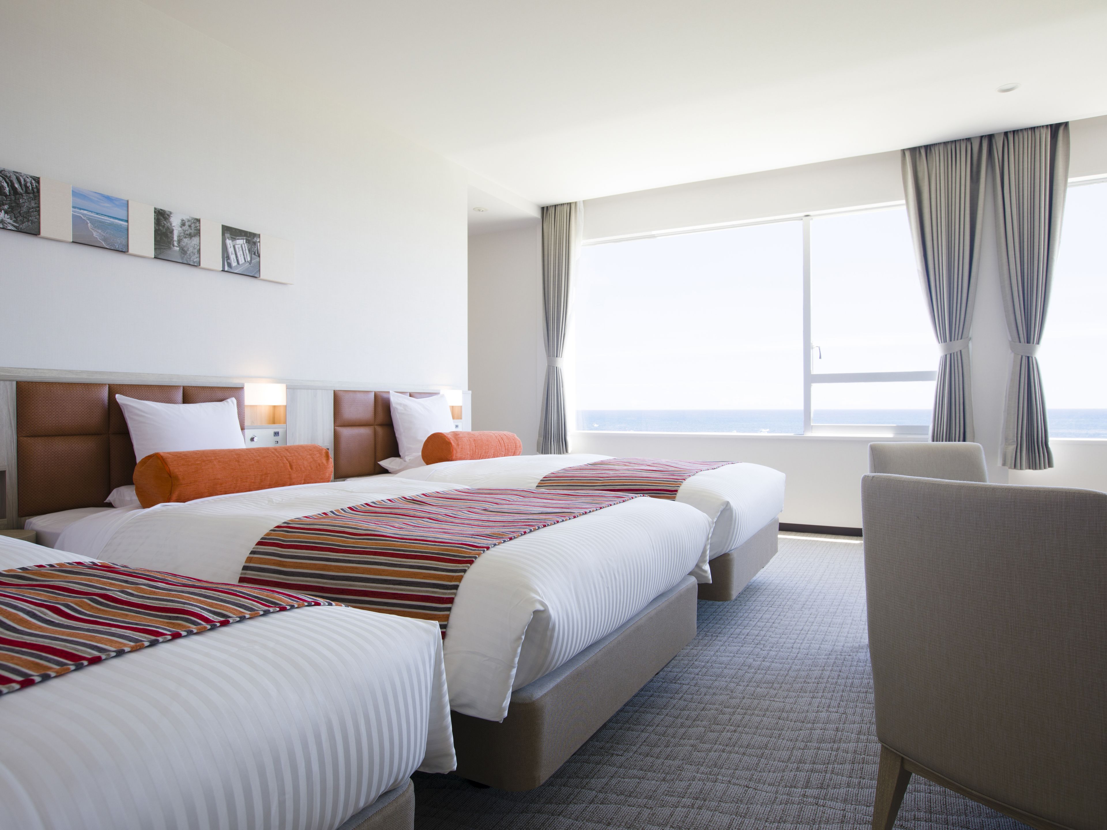 A hotel room with two beds, a large window with curtains, a chair, and artwork on the wall.