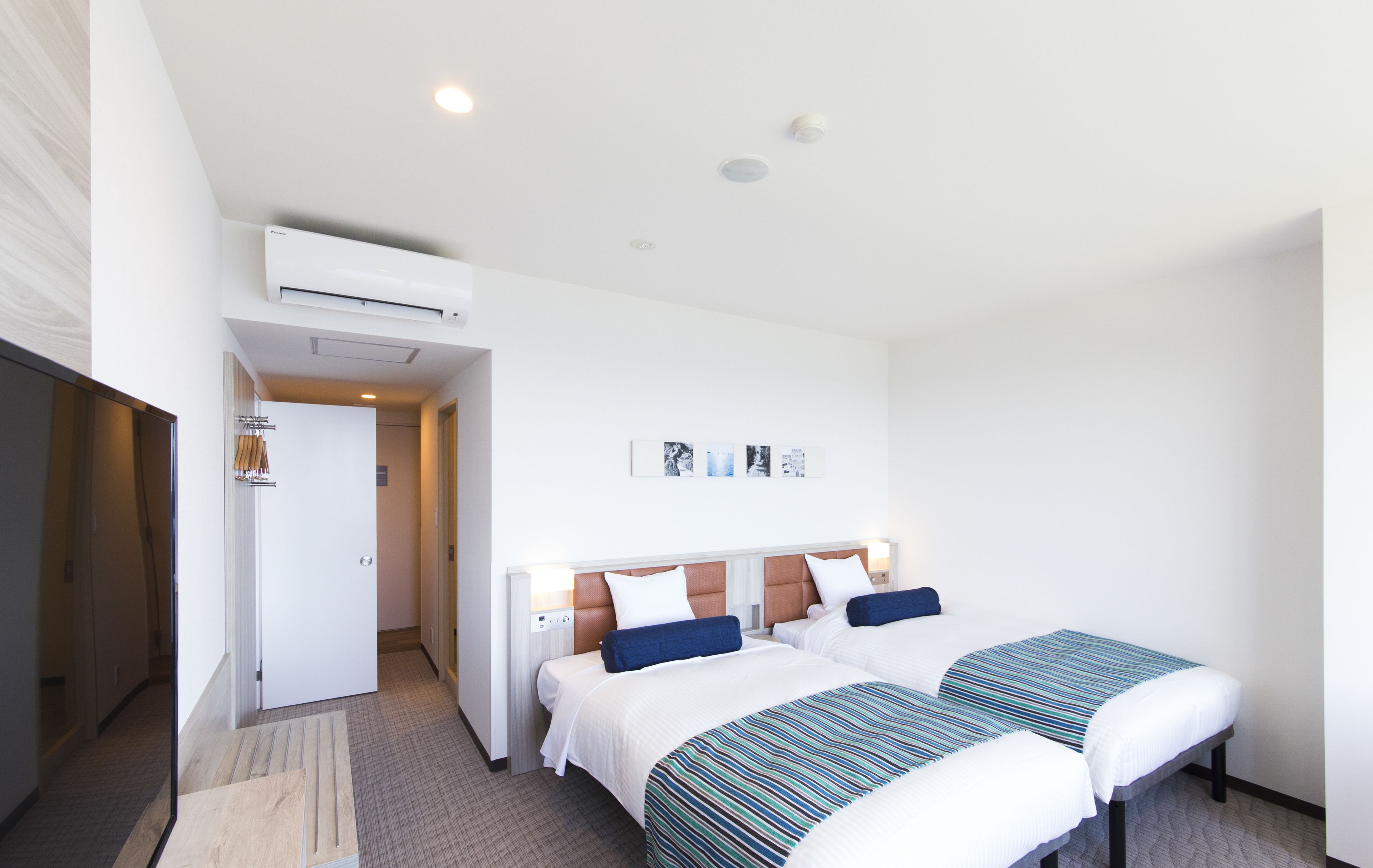 A hotel room with two beds, a white air conditioning unit, a door leading to another room, and a small table with a striped tablecloth.