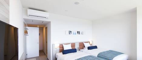 Ein Hotelzimmer mit zwei Betten, einer weißen Klimaanlage, einer Tür zu einem weiteren Raum und einem kleinen Tisch mit gestreifter Tischdecke.