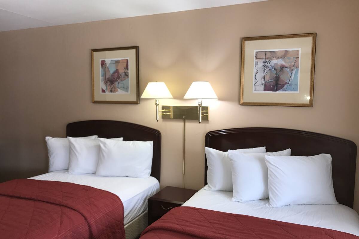 A hotel room with two beds, each with white bedding and red throw blankets. There are two wall-mounted lamps between the beds, and two framed pictures above them.