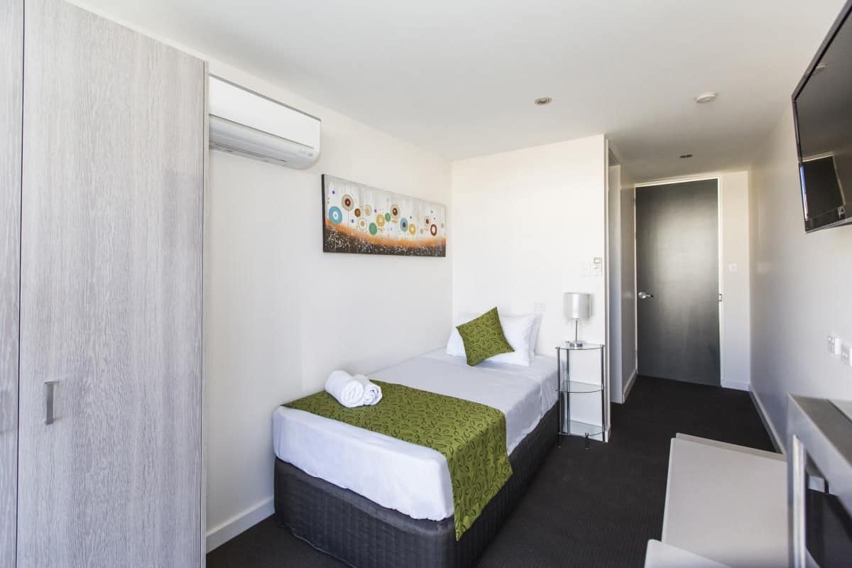 Quarto de hotel com cama, mesa de cabeceira com abajur, um quadro na parede e um guarda-roupa.