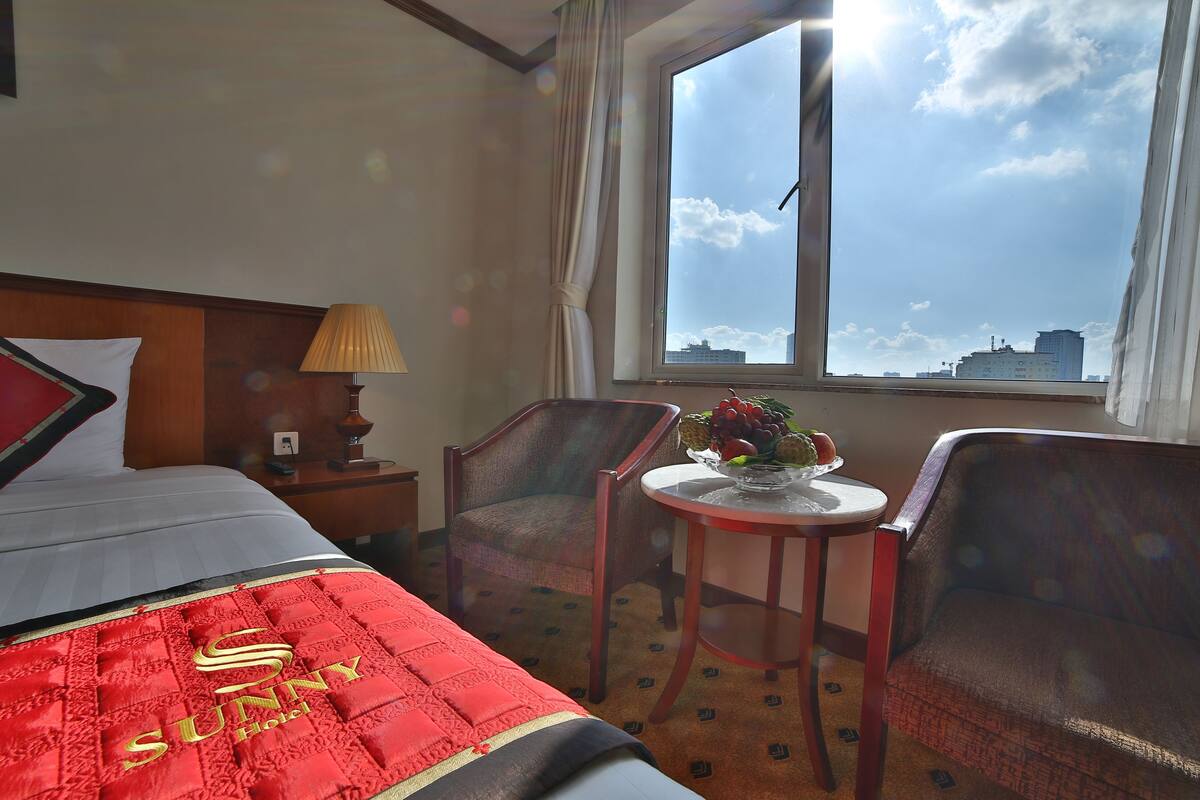 Une chambre d’hôtel avec un lit, deux fauteuils, une petite table avec des fruits, une lampe et une fenêtre donnant sur la ville.