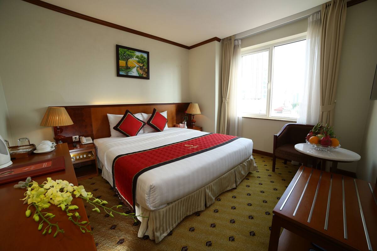 Une chambre d’hôtel avec un lit, des tables de chevet, un bureau, une chaise, une petite table avec des fruits et une fenêtre avec des rideaux.
