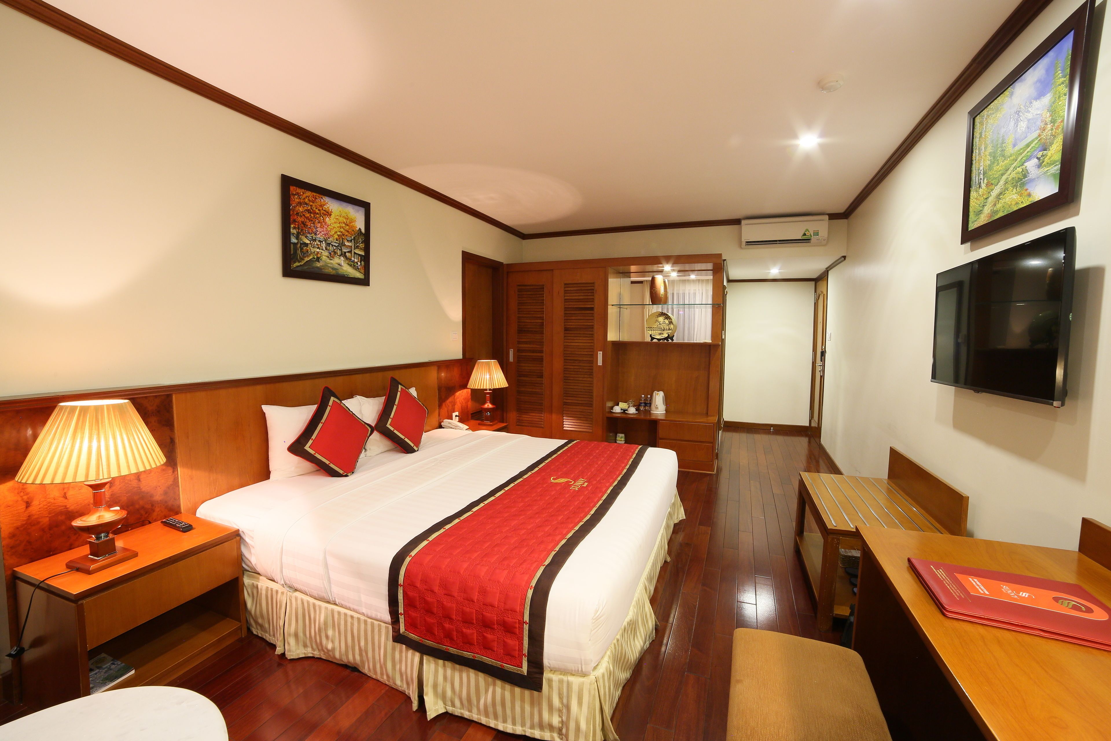 A hotel room with a bed, bedside lamps, a TV, a wooden table, and a desk.